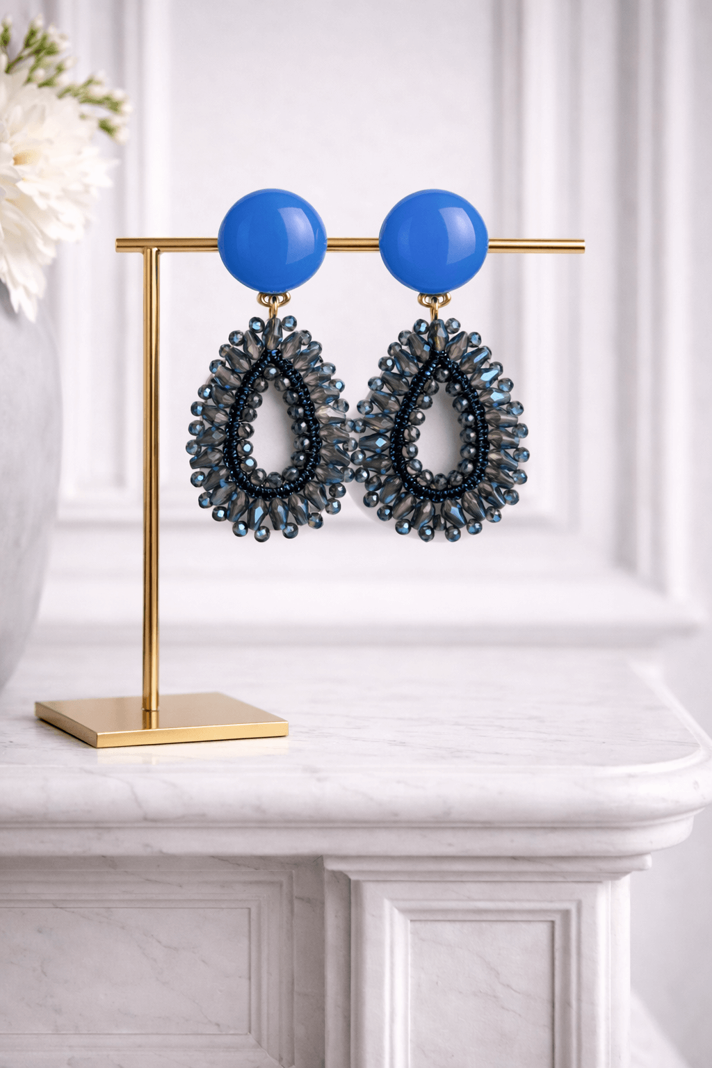 Blaue Garne-Ohrringe mit Glasperlen in Tropfenform an goldfarbenem Schmuckständer auf hellem Marmortisch.

