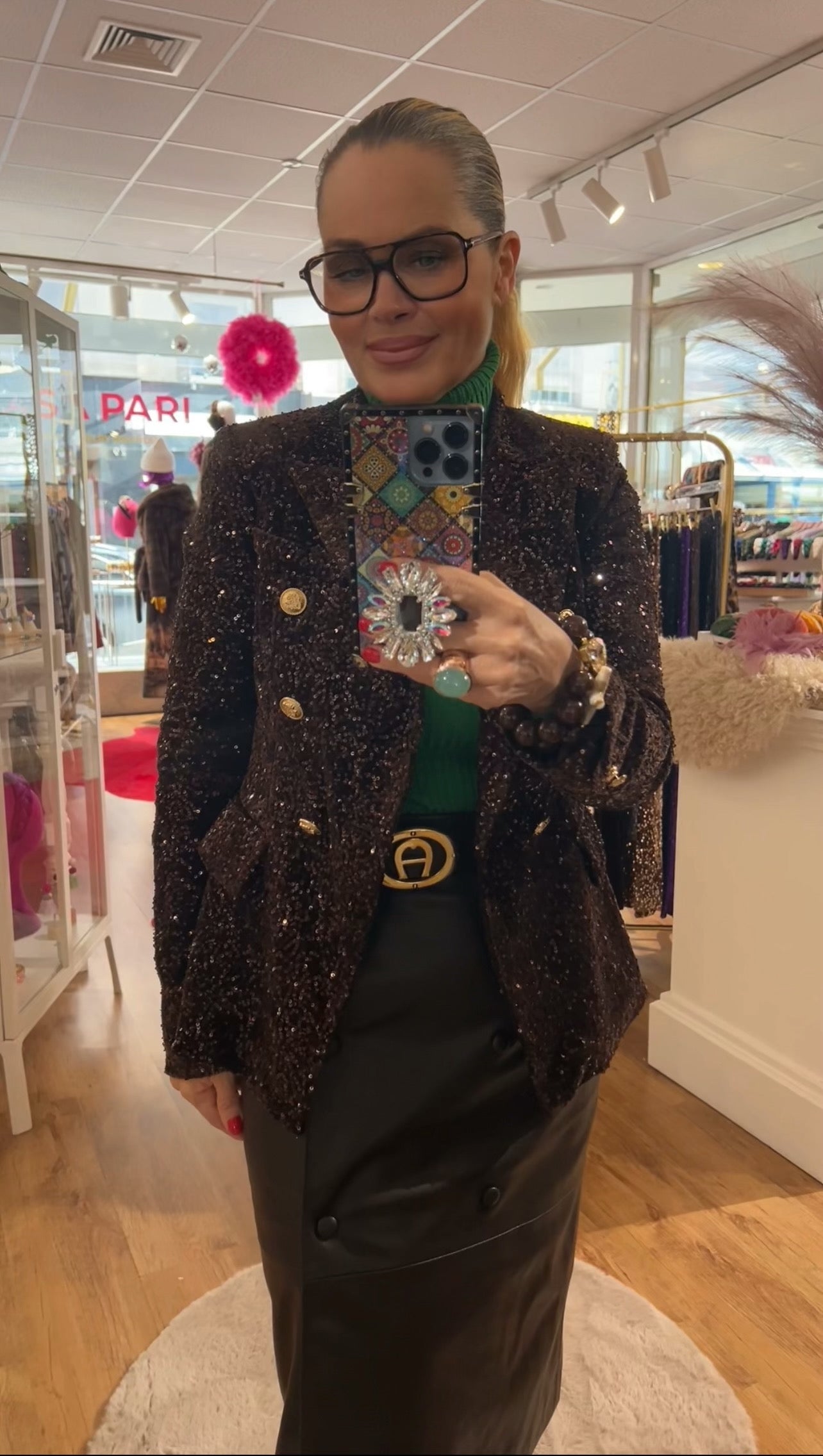 Frau steht im Laden vor einem Spiegel und macht ein Spiegel Selfie. Sie trägt einen dunklen schimmernden Pailletten Blazer mit goldfarbenen Knöpfen, einen grünen Rollkragenpulli und einen schwarzen Rock. In der Hand hält sie ein Smartphone mit gemustertem Case und großen Ring. Im Hintergrund eine Ladeneinrichtung.