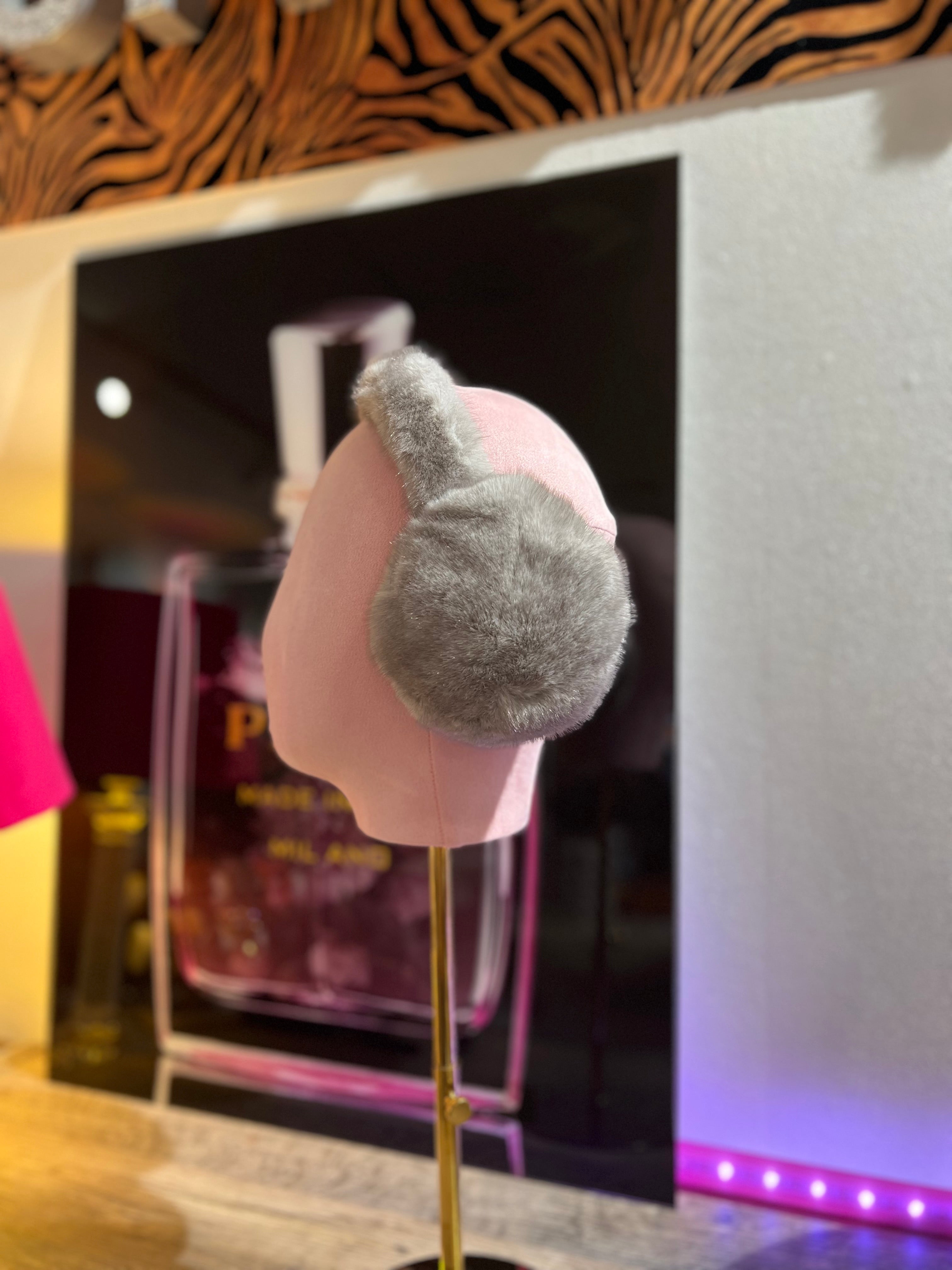 Hellgrau, flauschige Ohrwärmer auf rosafarbene Präsentation Büste, im Hintergrund unscharf ein Parfumflakon und warme Beleuchtung.