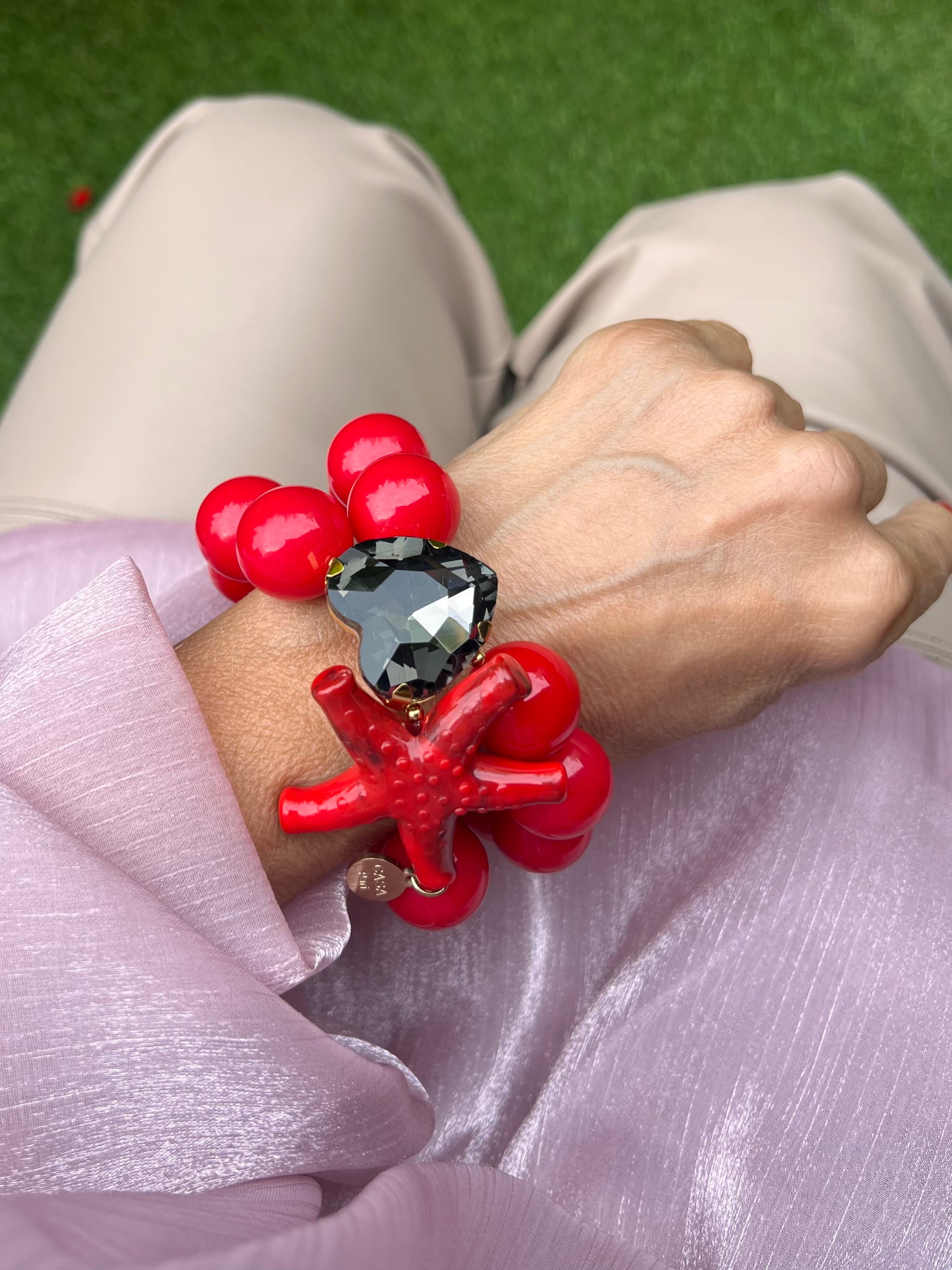 Armband mit zwei Reihen glänzender roten Perlen, verziert mit einem funkelndem Kristalle Herz in Grau, auf hellem Hintergrund.