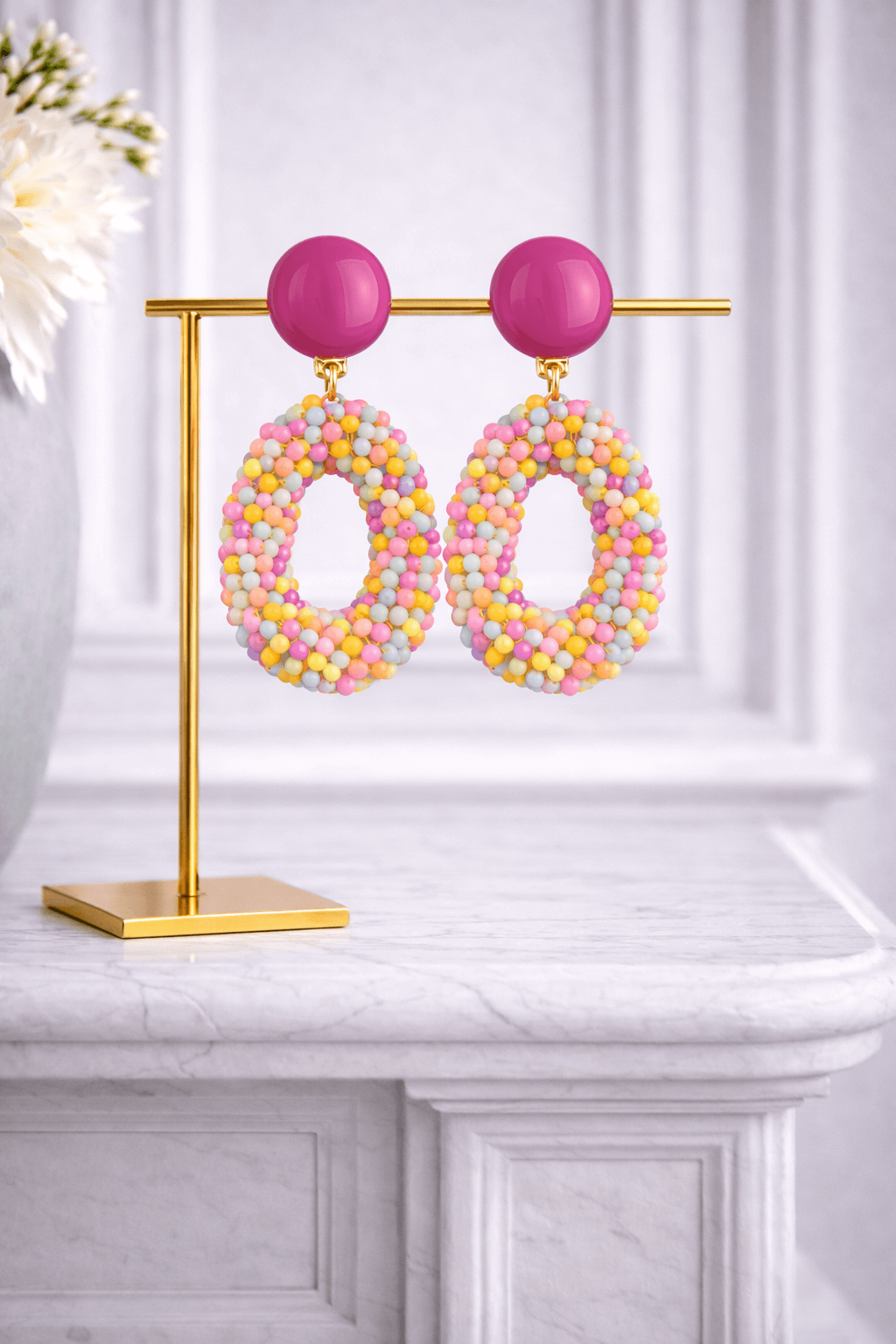Ovale Ohrringe mit pinkem Clip-Verschluss und bunten Perlen in Pastelltönen, präsentiert auf goldenem Schmuckständer vor hellem Hintergrund.
