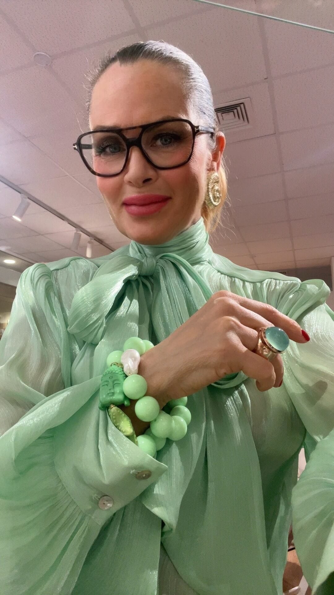 Person trägt eine mintgrüne, transparente Bluse mit Schleifenkragen, dazu ein Armband aus mintgrünen Perlen mit grünem Buddha-Element, weißer Perle und kleinem Seestern-Anhänger am Handgelenk. Ergänzt durch eine grüne Ringfassung mit rundem Stein und goldfarbene Ohrringe, aufgenommen in einem Innenraum.