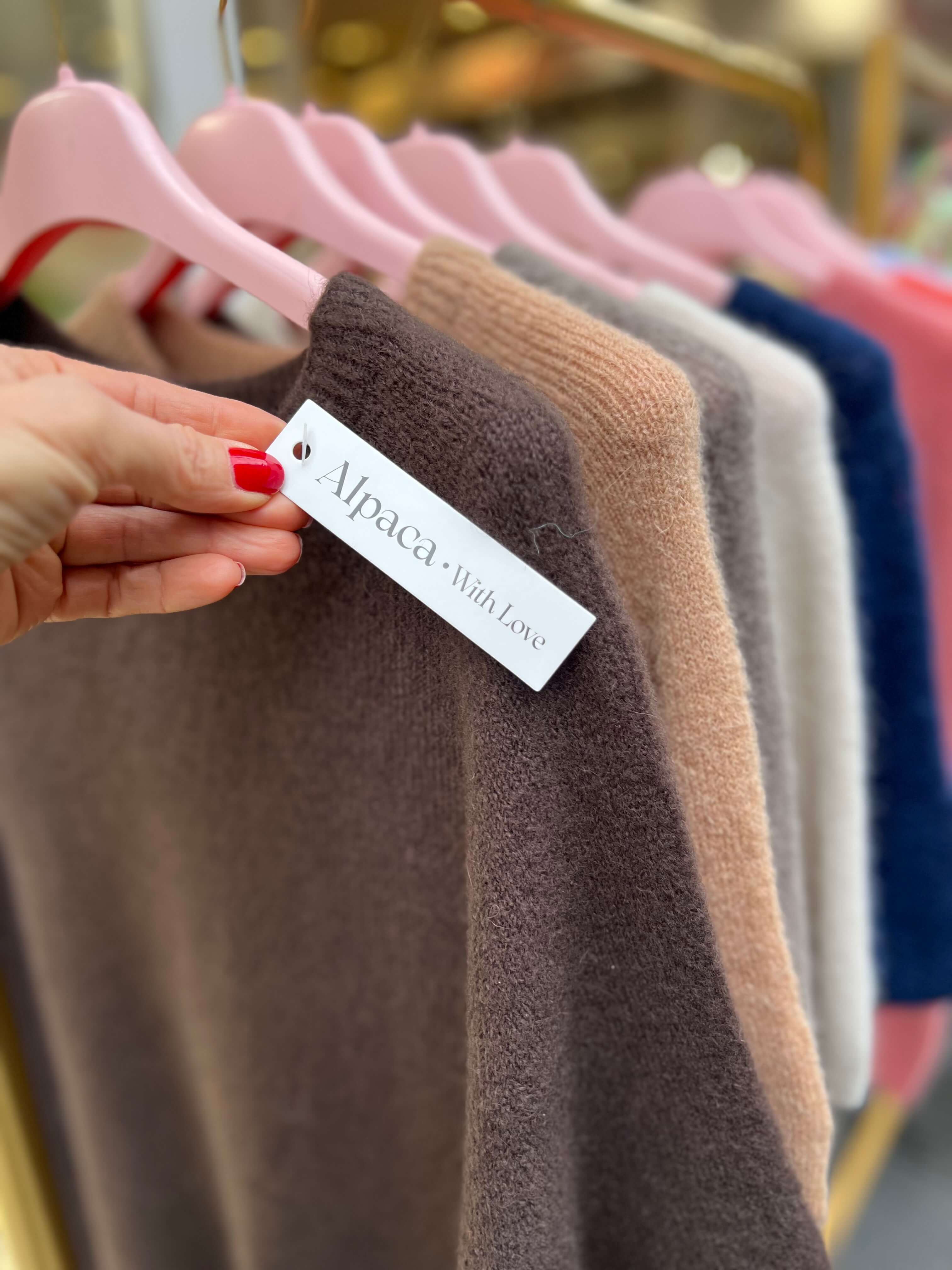 Nahaufnahme eines kurzärmeligen Strickpullovers in Braun aus Alpaka-Mischung auf einem rosa Kleiderbügel. Eine Hand mit rot lackierten Fingernägeln hält ein weißes Etikett mit der Aufschrift „Alpaca. With Love“. Im Hintergrund hängen weitere Pullover in Beige, Grau, Creme, Dunkelblau und Rosa unscharf an einer goldfarbenen Kleiderstange.