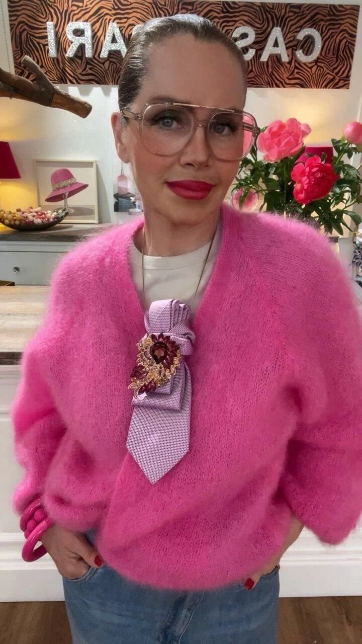 Frau trägt einen pinken Kid-Mohair-Cardigan mit Jeans und dekorativem Accessoire im Brustbereich, frontal in einer Boutique.