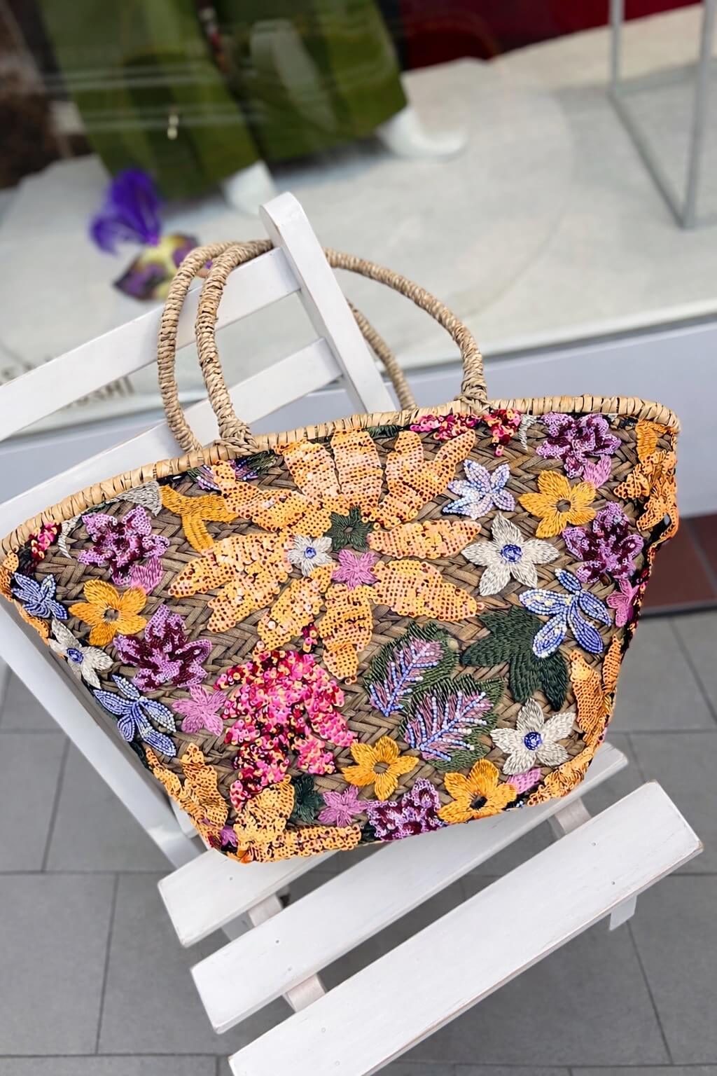 Geflochtene Basttasche mit zwei runden Henkeln, überzogen mit transparentem Stoff und dicht bestickt mit schimmernden Pailletten in warmem Gelb, Pink, Lavendel, Creme und Grüntönen in floralen Mustern.