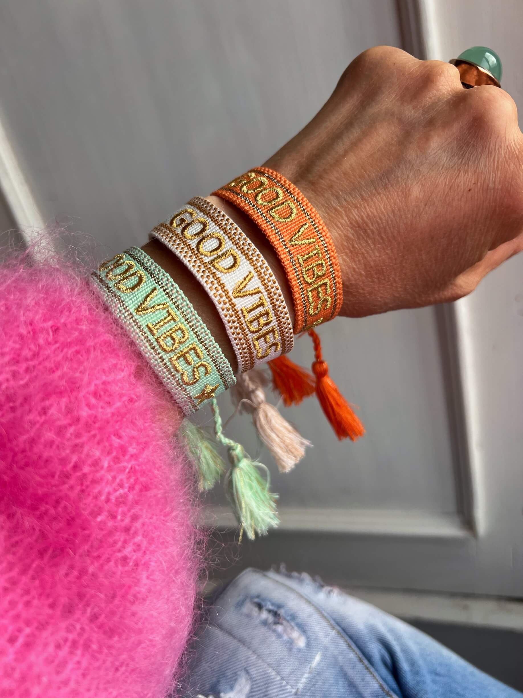 Gewebte Stoffarmbänder mit der Aufschrift „GOOD VIBES“ in Goldschrift, getragen am Handgelenk.
Farben: Mintgrün, Creme und Orange.
Jedes Armband mit seitlichen Quasten.
Person trägt einen pinkfarbenen, flauschigen Pullover und eine Jeans.
Auf dem Finger ein Ring mit grünem Stein.