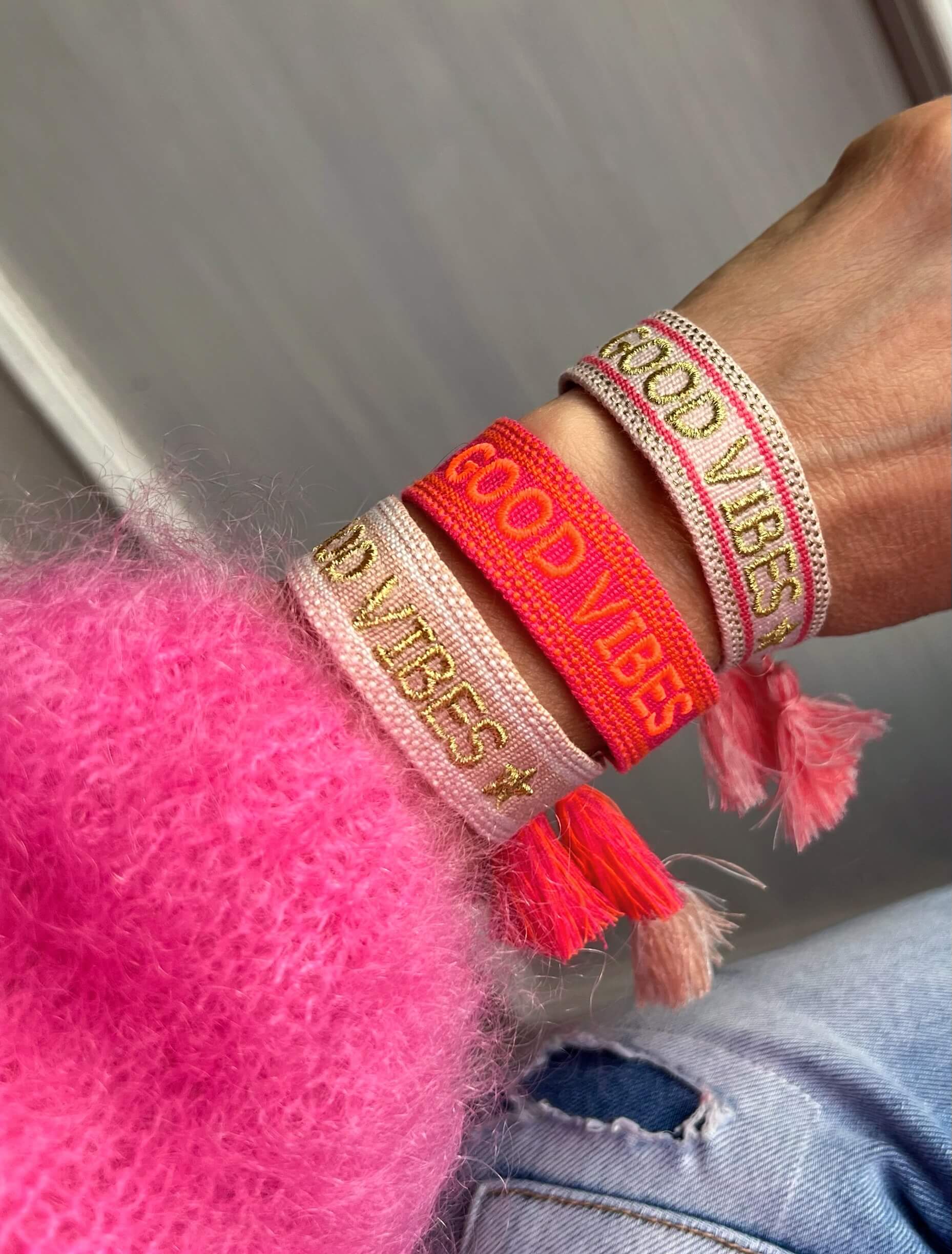 Handgelenk mit drei gewebten Stoffarmbändern in Pink-, Beige- und Koralltönen mit goldfarbener Schrift „GOOD VIBES“ und Quasten; getragen zu pinkem Pullover und heller Jeans.