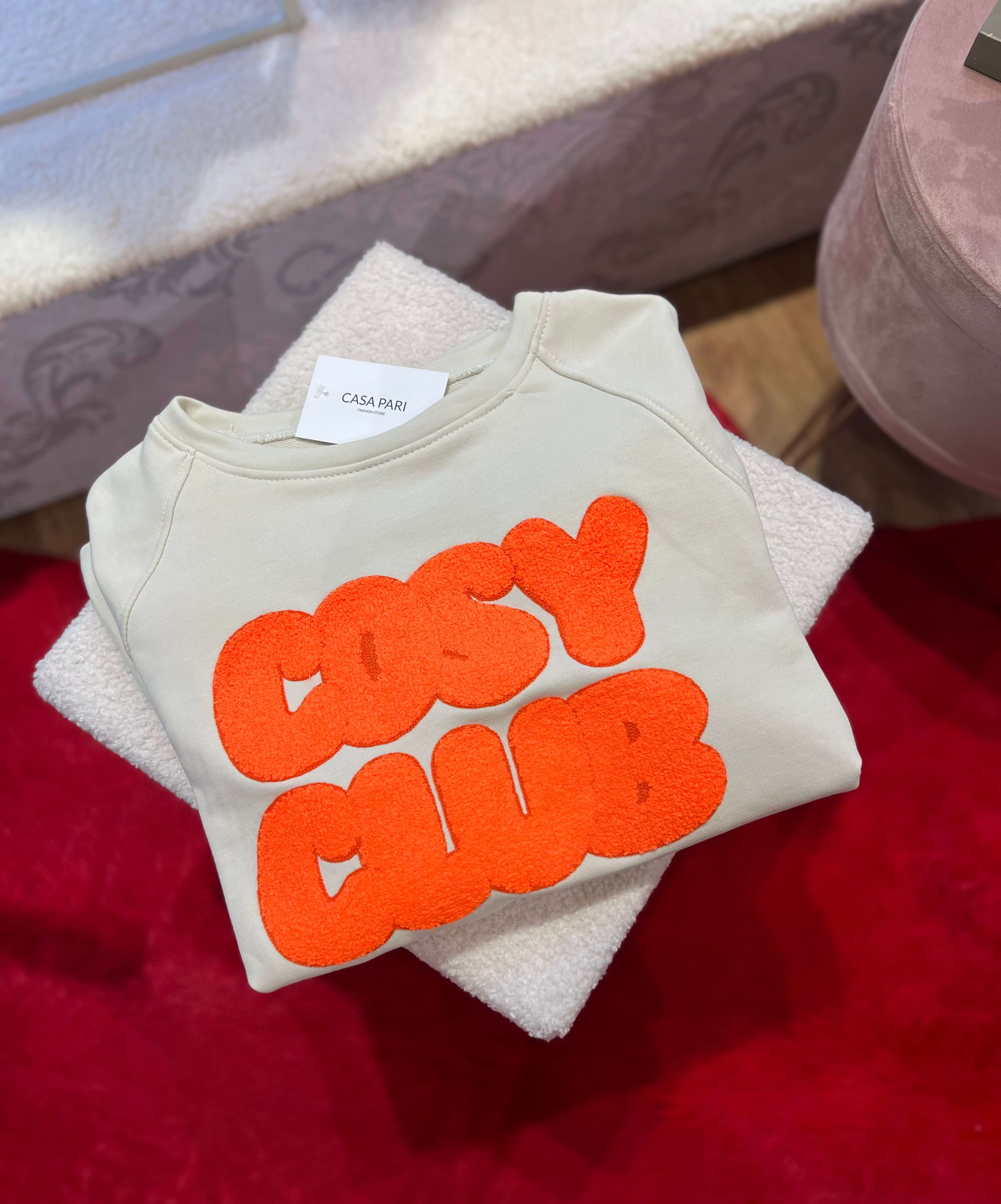 Gefaltetes, hellbeiges Sweatshirt mit rundem Halsausschnitt und großem, flauschigem Schriftzug „Cozy Club“ in Neon-Orange auf der Vorderseite.

Das Sweatshirt liegt auf einer hellen, strukturierten Unterlage; darunter ist ein roter Teppich zu sehen. Oben am Kragen befindet sich ein weißes Etikett mit der Aufschrift „CASA PARI“.