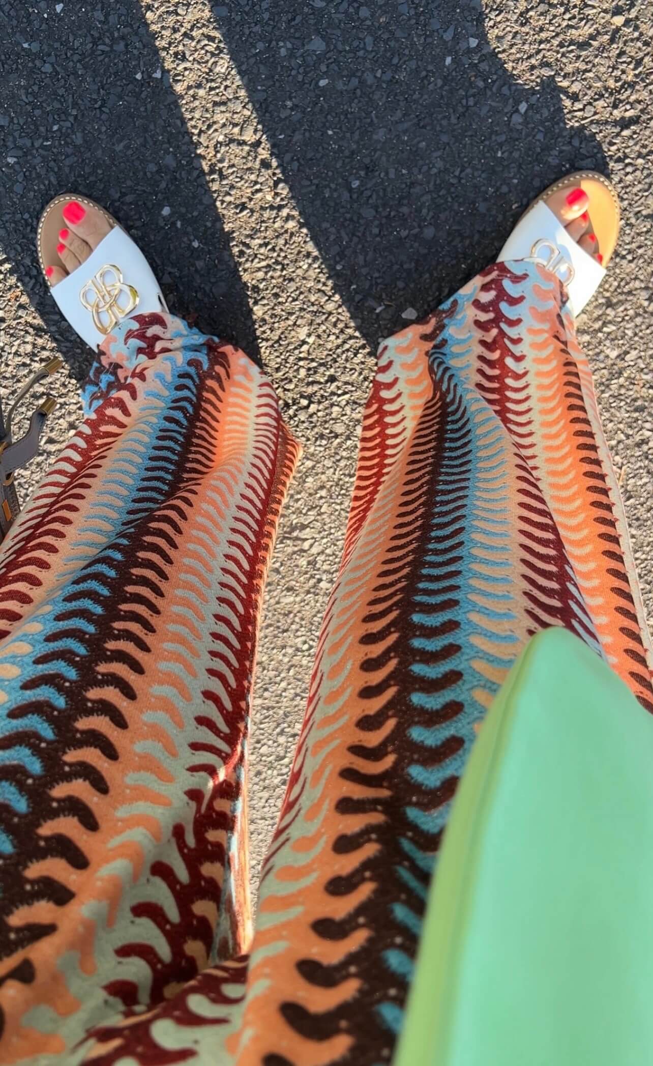 Detailaufnahme einer gemusterten Palazzo-Hose in Türkis, Peach und Bordeaux mit weitem Bein, kombiniert mit weißen Sandalen und Tasche, fotografiert im Sonnenlicht auf Asphalt.