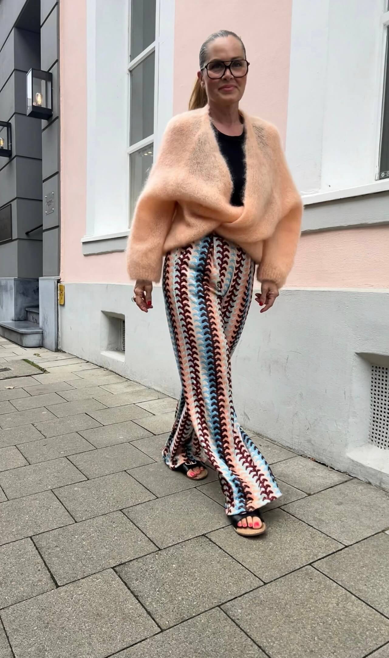 Person trägt gemusterte Palazzo-Hose in Türkis, Peach und Bordeaux mit weitem Bein, kombiniert mit apricotfarbenem Strickpullover, fotografiert auf einer Straße vor einem Gebäude.