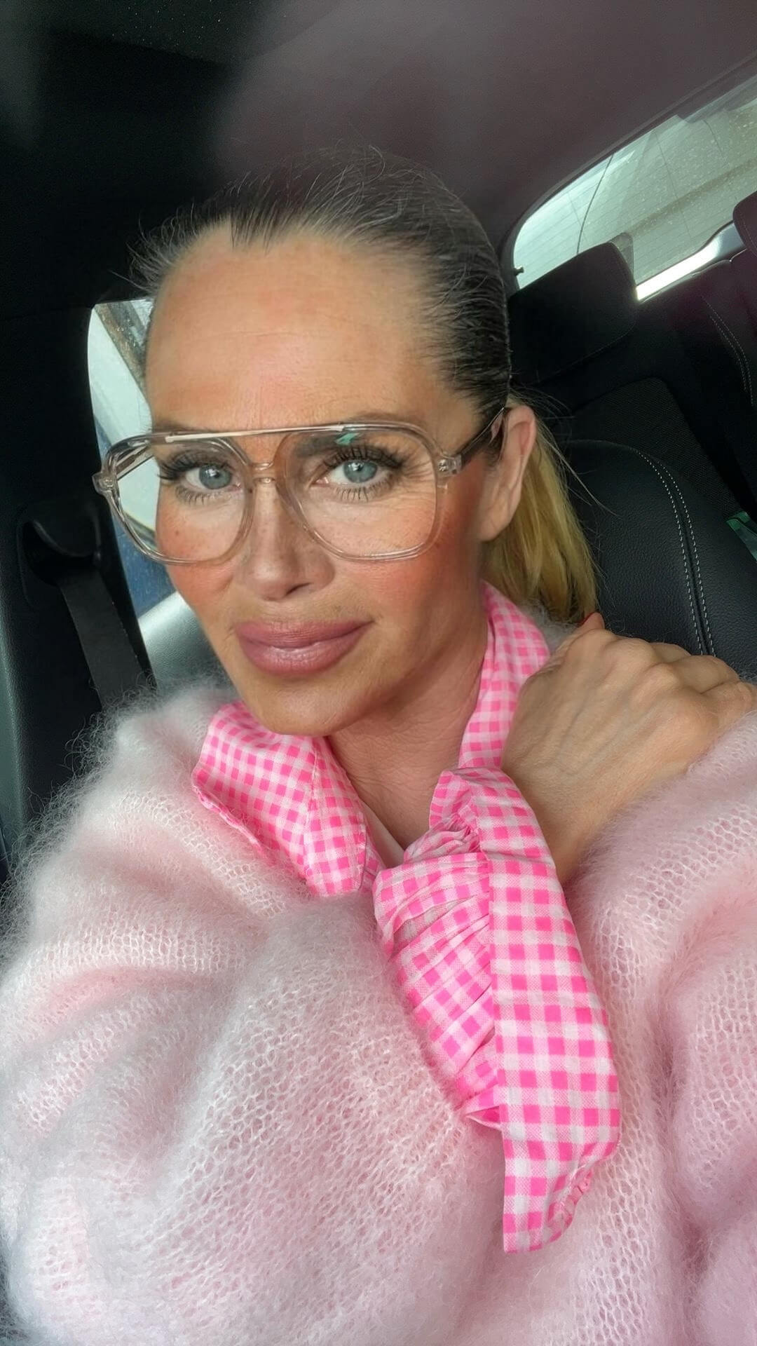 Frau mit zurückgebundenem Haar sitzt im Auto und trägt eine transparente Brille, ein rosa Mohair-Strickoberteil und ein pink-weiß kariertes Hemd mit Schleife am Hals. Eine Hand liegt auf der Schulter.
