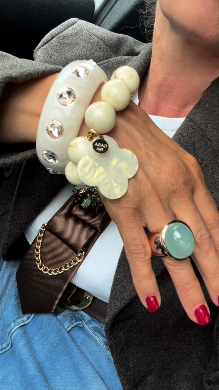 Handgelenk mit beigen Resina-Armreif mit Strass-Steinen, kombiniert mit einem cremefarbenen Perlenarmband und einem grünen Ring. Die Person trägt einen grauen Blazer, Jeans und eine braune Krawatte mit goldfarbener Kette.