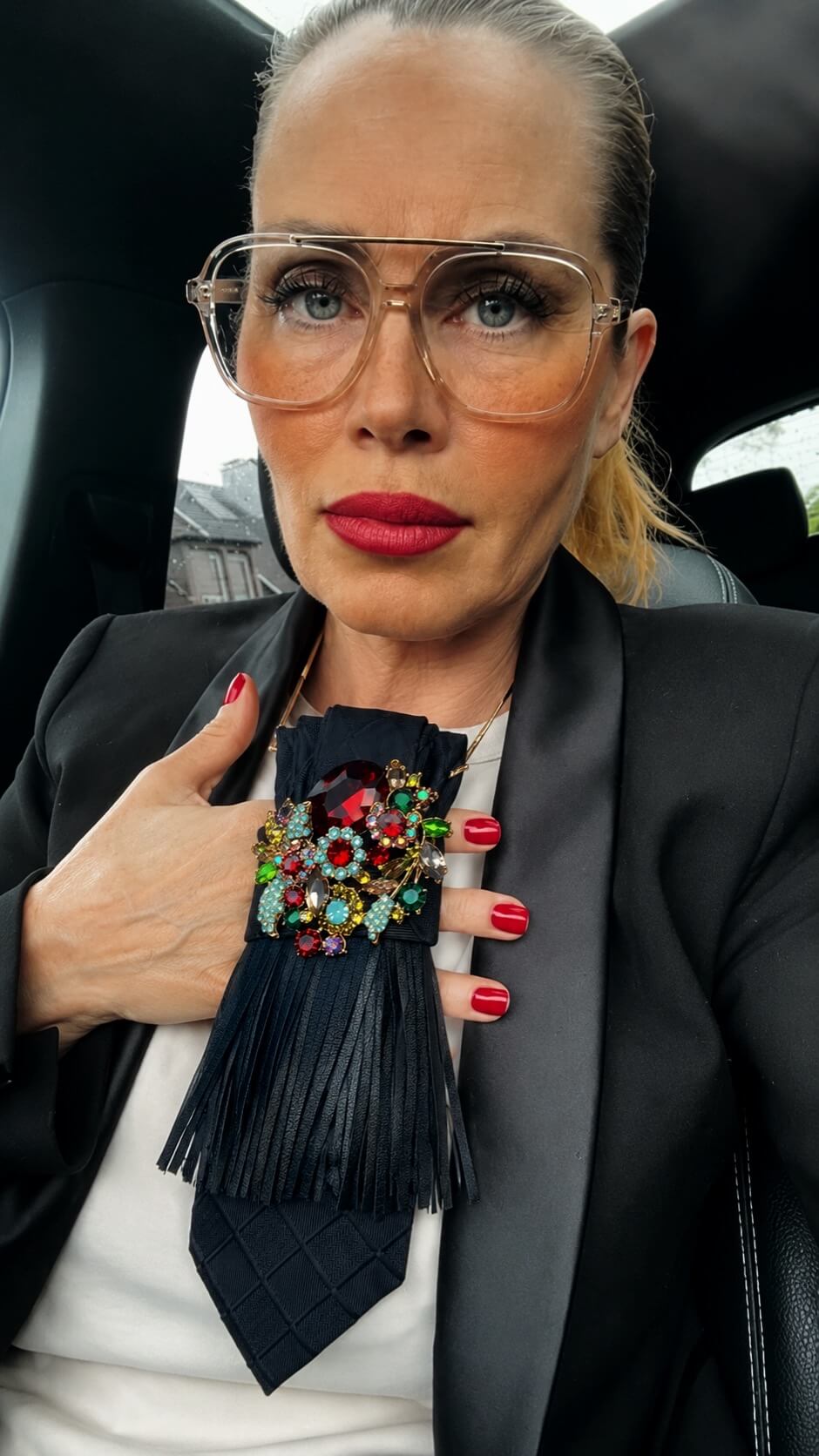 Frau mit heller Bluse, schwarzem Blazer und großer transparenter Brille trägt eine schwarze Statement-Krawatte mit Lederfransen, goldfarbener Kette und großer Schmuckbrosche mit roten, grünen und türkisfarbenen Steinen. Die Aufnahme entstand im Innenraum eines Fahrzeugs.