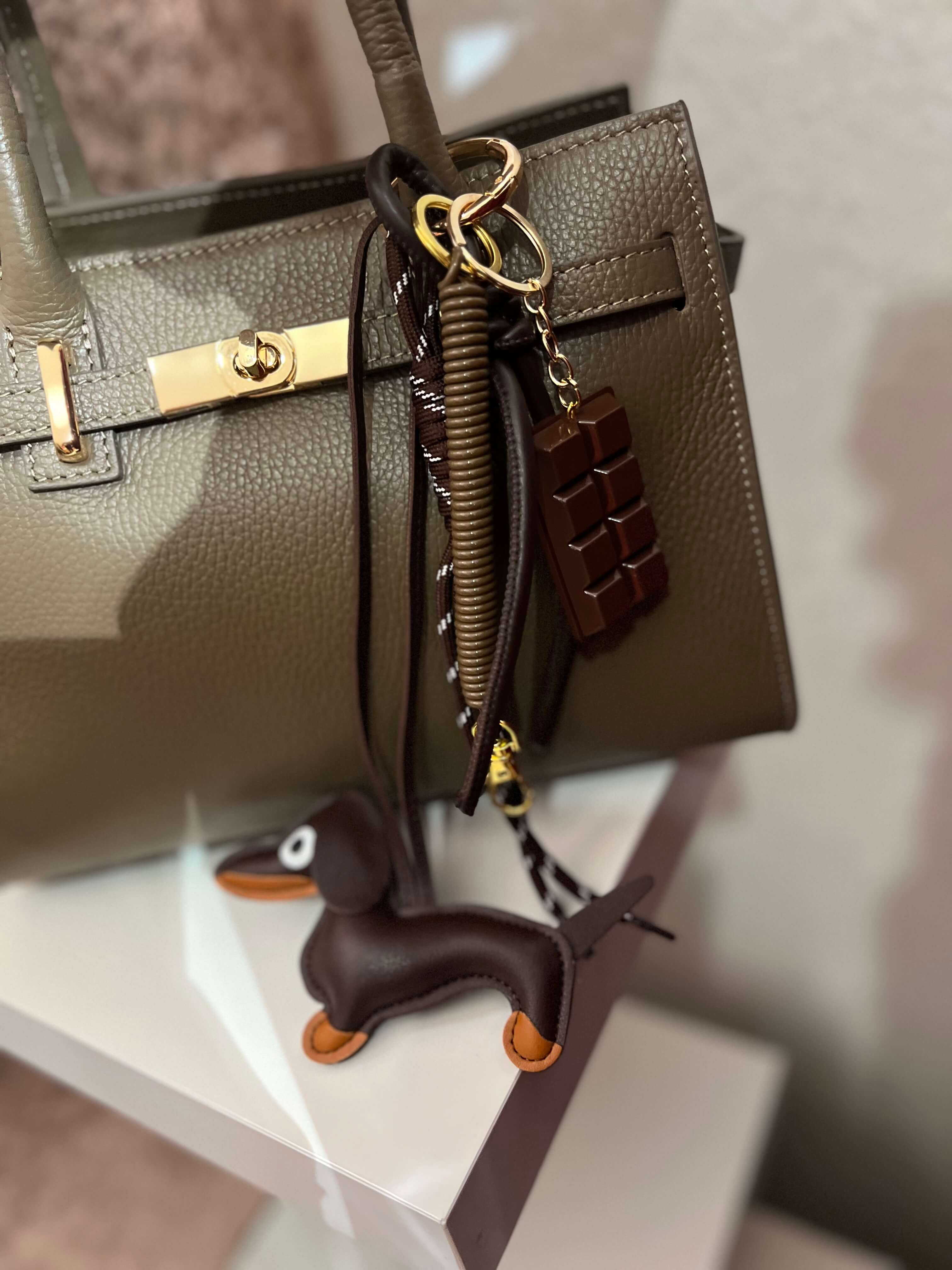 Taschenanhänger in Form eines braunen Dackels aus Kunstleder, befestigt an einer olivfarbenen Handtasche mit goldfarbenem Clip und zusätzlichen dekorativen Anhängern.