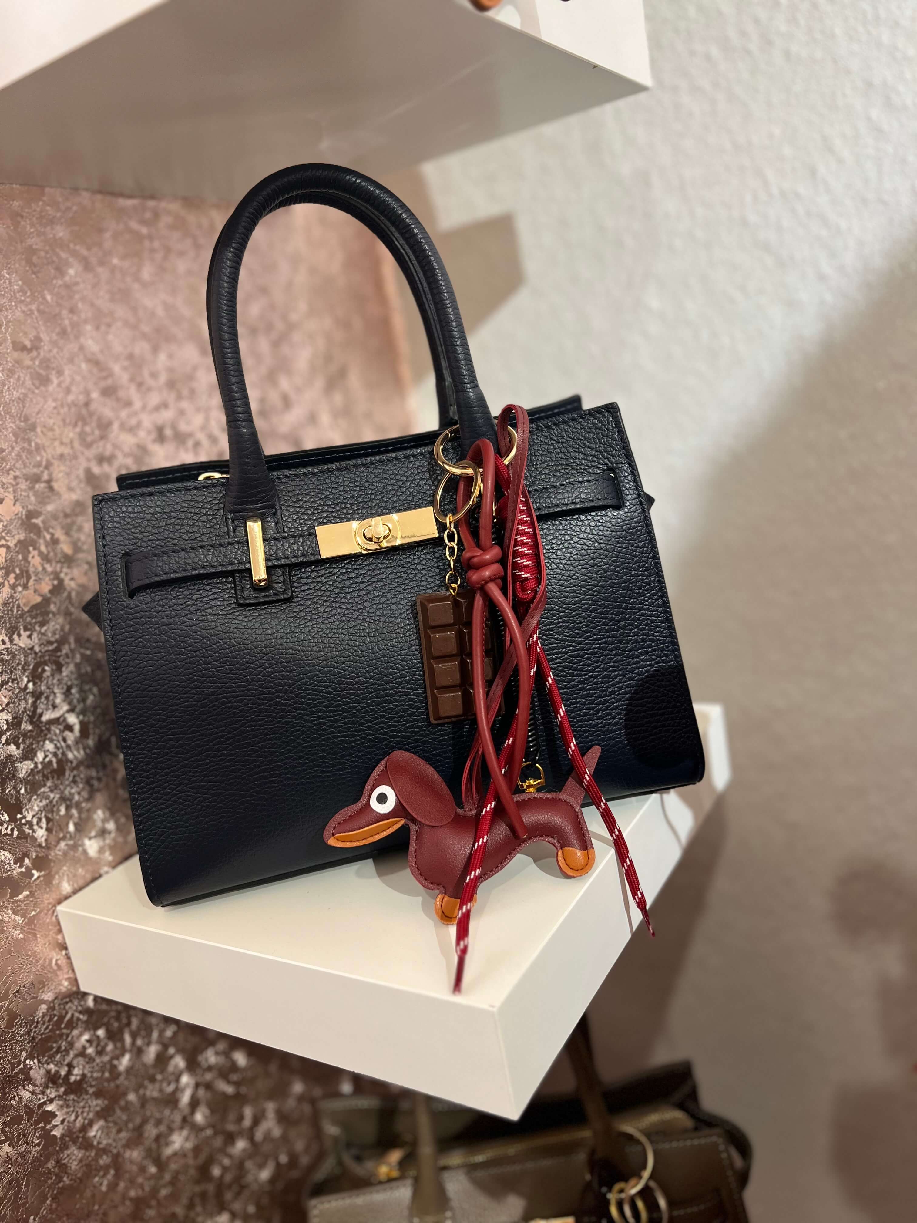 Taschenanhänger in Form eines braunen Dackels aus Kunstleder, befestigt an einer blauen Handtasche mit goldfarbenem Clip und zusätzlichen dekorativen Anhängern.