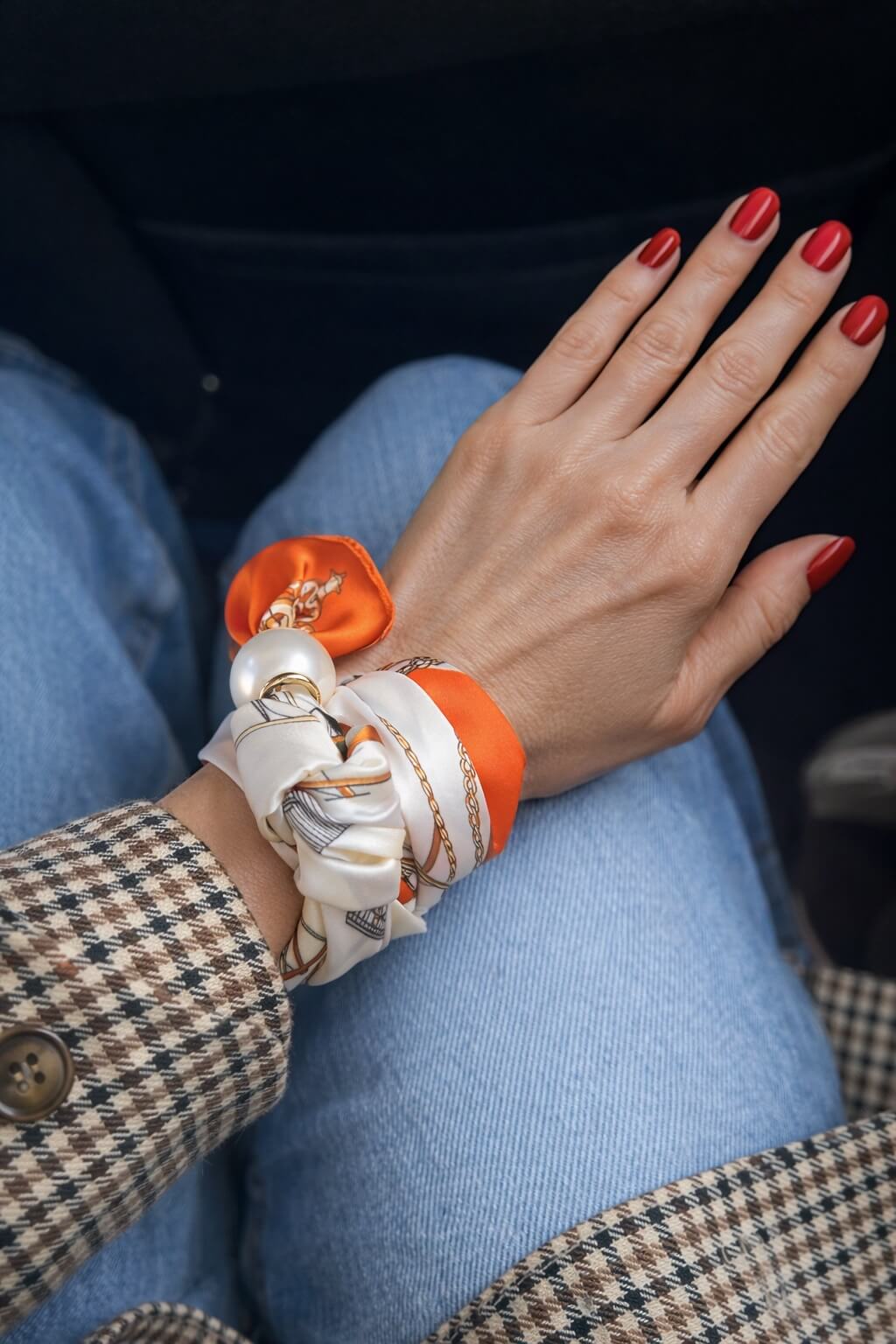 Hand mit rotem Nagellack trägt ein orange-weißes Seidentuch mit Perle am Handgelenk im Auto