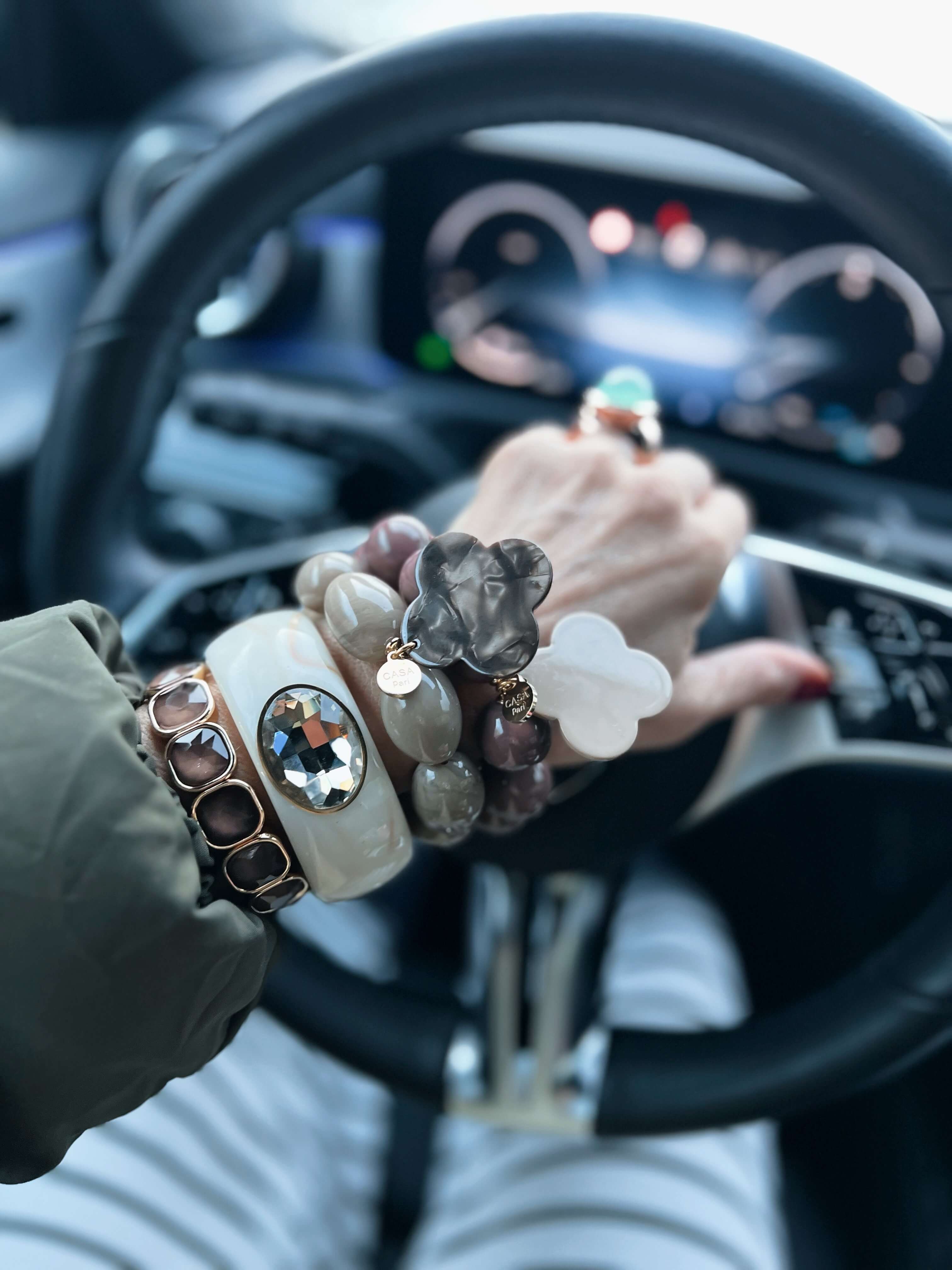 Handgelenk mit mehreren Armbändern in Creme-, Taupe- und Brauntönen, darunter runde und ovale Kunstharz-Perlen sowie Kleeblatt-Anhänger. Aufgenommen im Auto, Hand am Lenkrad, unscharfer Fahrzeug-Innenraum im Hintergrund.