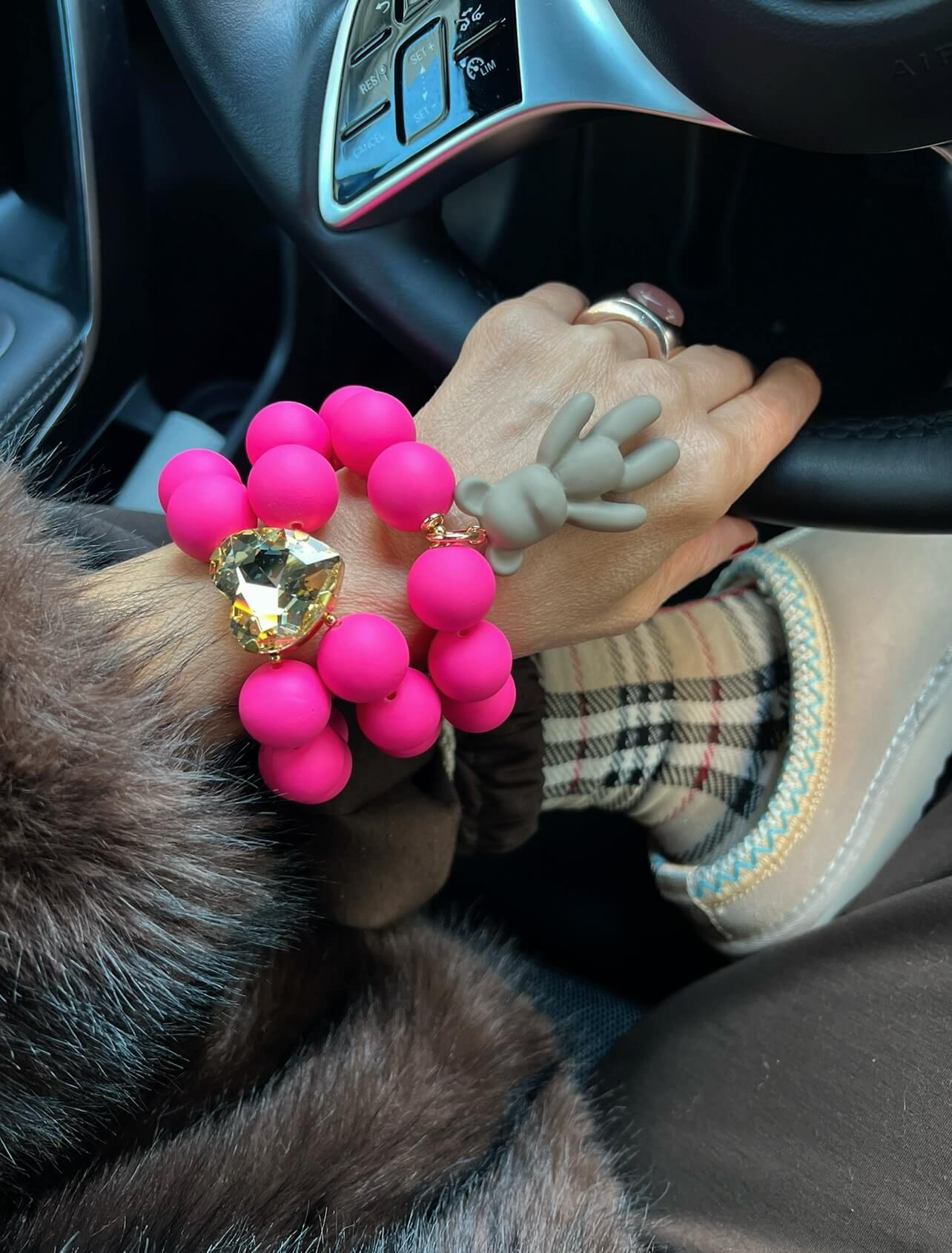 Handgelenk mit zwei pinken Armbändern mit großen runden Perlen, einem goldfarbenen Herzanhänger und einem grauen Bunny-Anhänger, aufgenommen im Auto vor einem Lenkrad.
