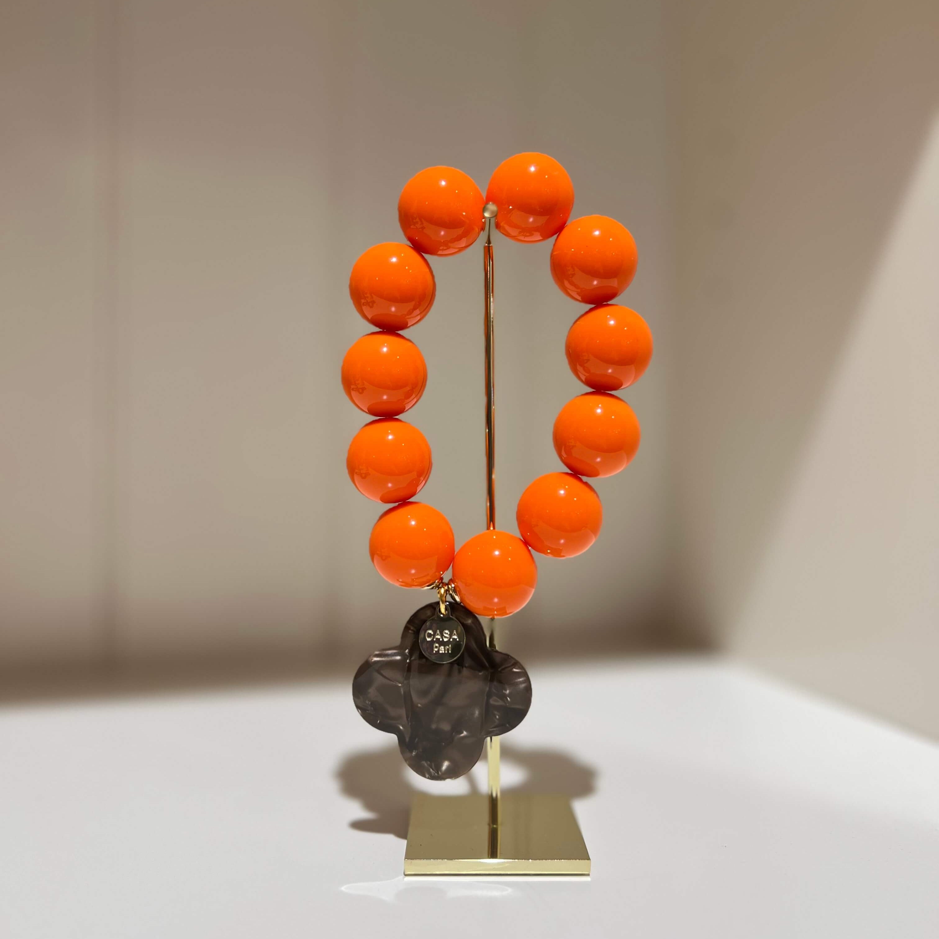 Orangefarbenes Armband mit runden Perlen und einem dunklen vierblättrigen Anhänger auf einem Schmuckständer.
