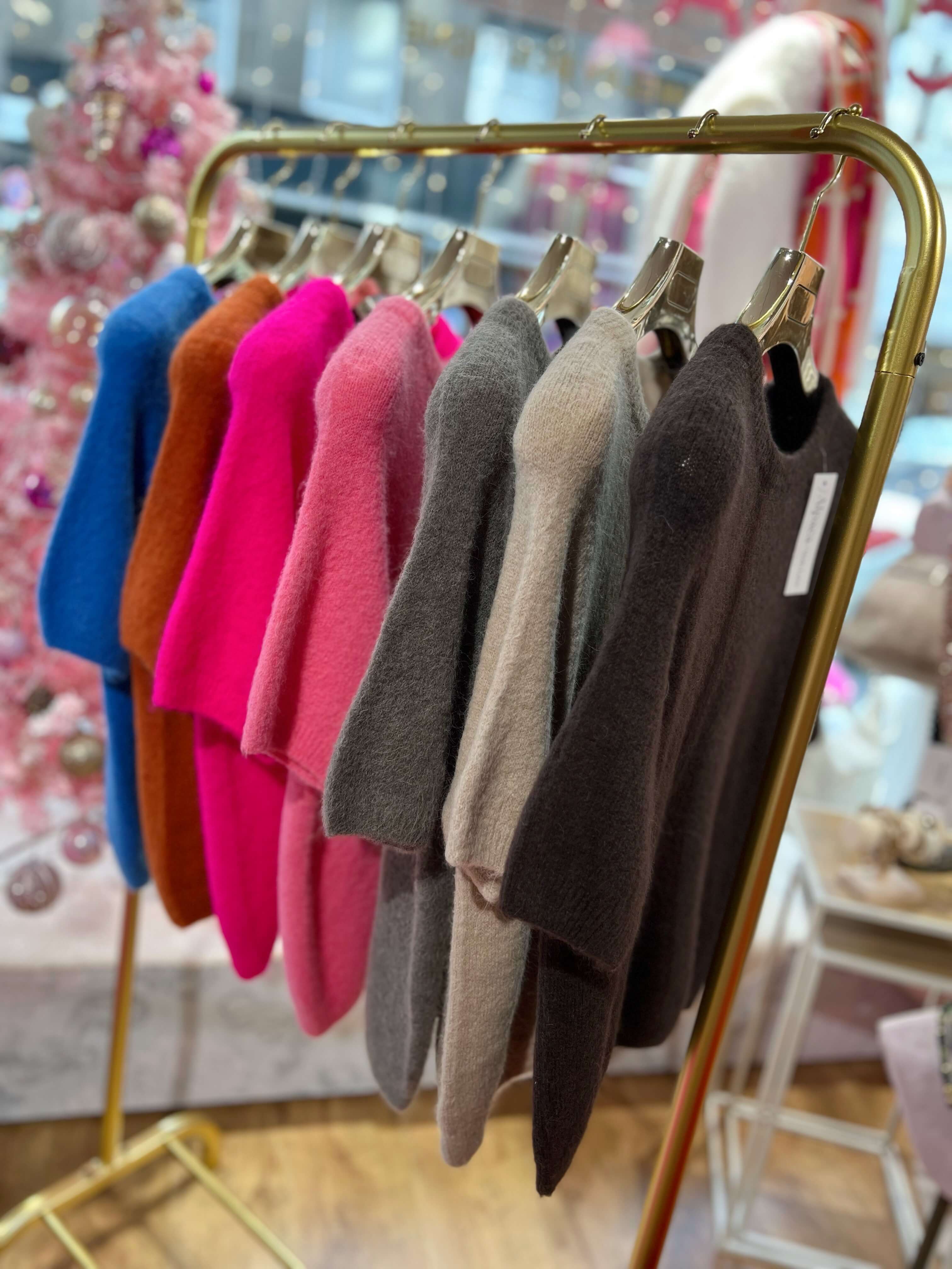 Mehrere kurzärmelige, flauschige Pullover in verschiedenen Farben hängen nebeneinander auf einer goldfarbenen Kleiderstange in einem Modegeschäft. Die Farben reichen von Blau, Braun und Pink über Rosé und Grau bis Beige und Dunkelgrau.