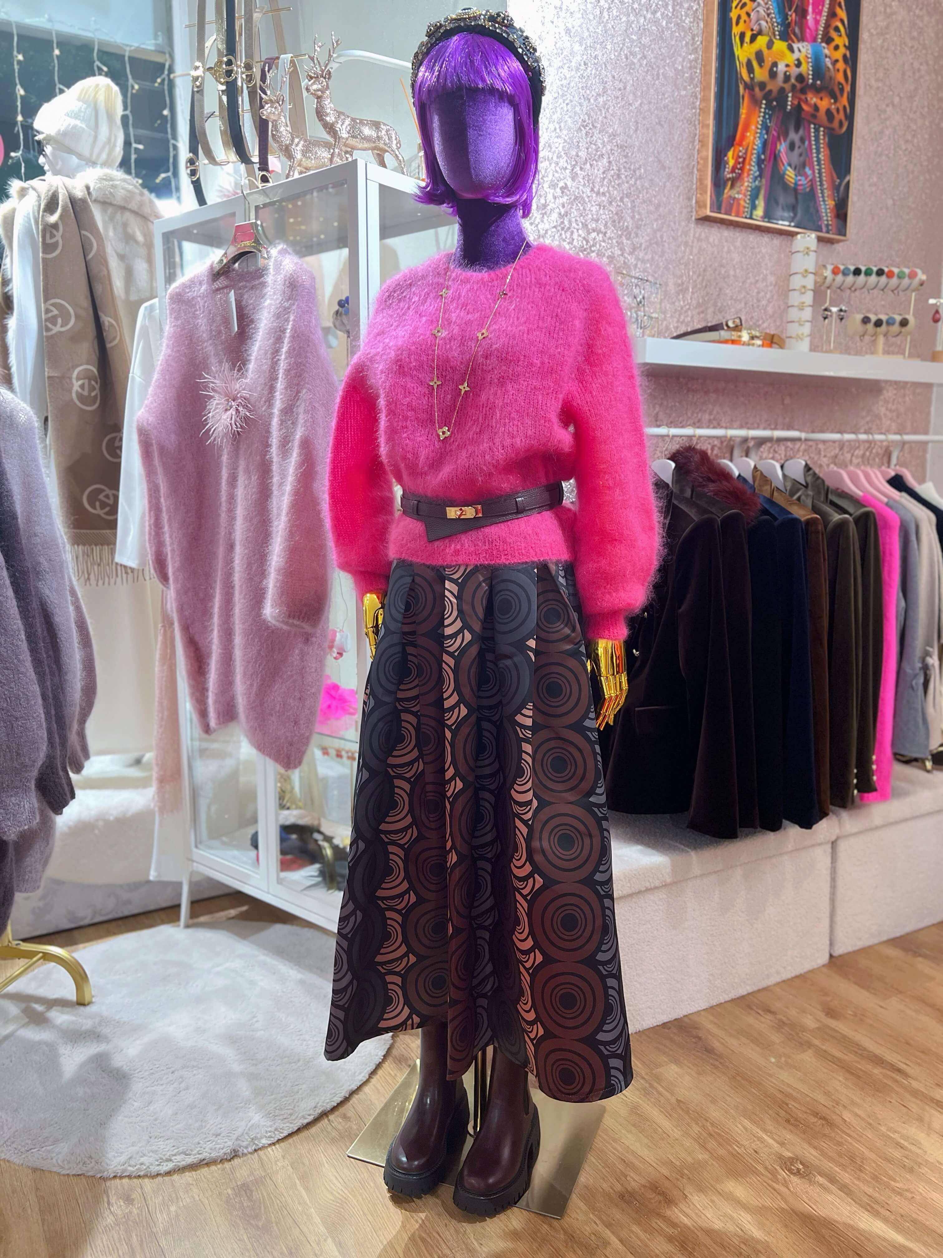 Schaufensterpuppe im Innenraum trägt einen pinken Strickpullover, einen knielangen Rock mit grafischem Kreis-Muster, Gürtel und dunkle Stiefel, umgeben von weiterer Kleidung.