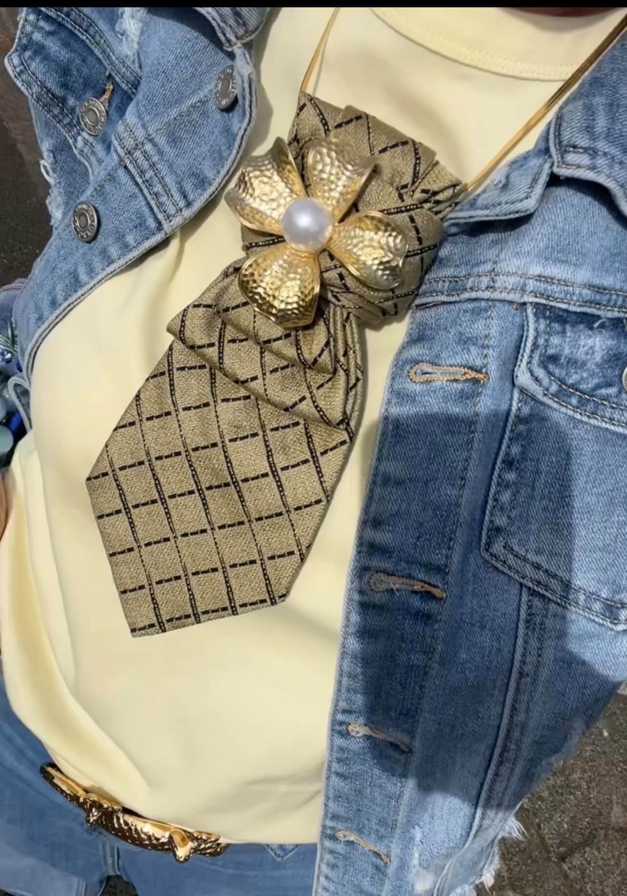 Frau trägt eine Vintage-Krawatte mit goldfarbener Brosche und feiner Kette, kombiniert mit dunkler Jacke, Nahaufnahme frontal vor neutralem Hintergrund
