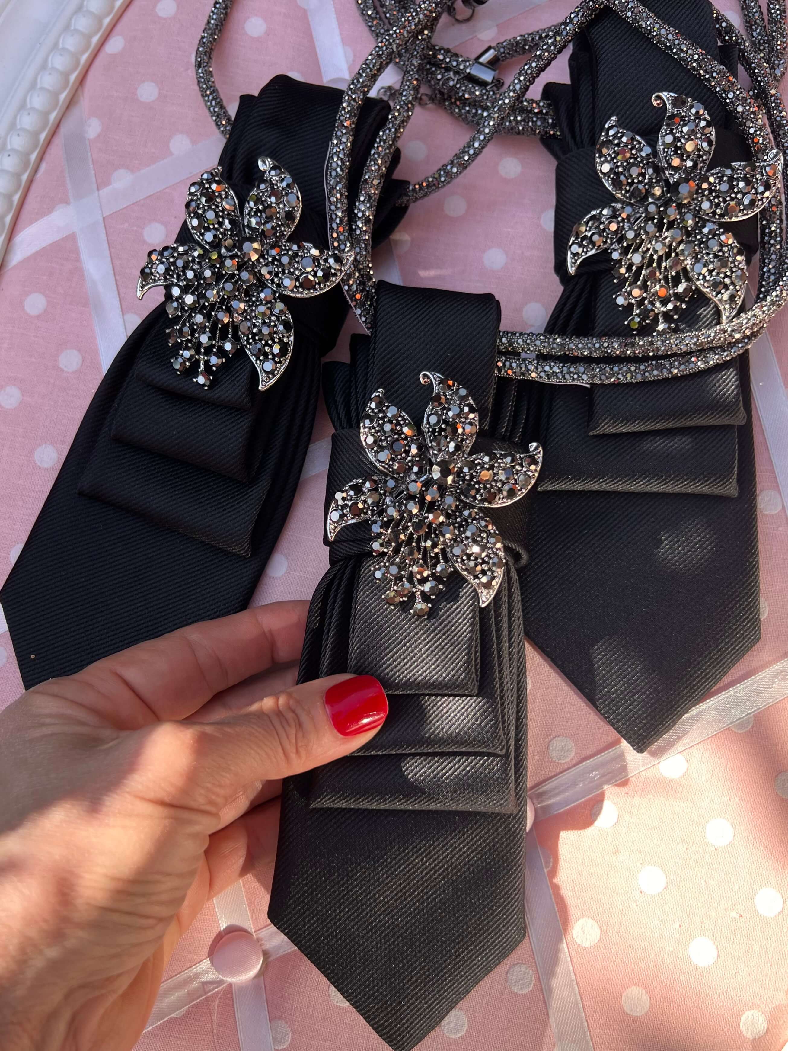 Schwarze Krawatten mit glatter Struktur, jeweils mit großer, funkelnder Blumenbrosche aus Strass verziert; mehrere Exemplare liegen auf rosa Untergrund mit weißen Punkten, eine Krawatte wird von einer Hand mit roten Fingernägeln gehalten.