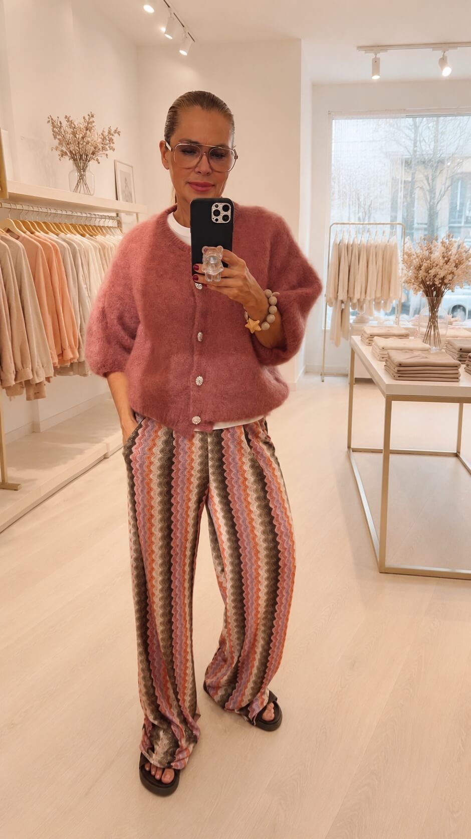 Frau steht in einer Boutique. Sie hat eine bunte Hose an mit einem Strick Cardigan und Strassknöpfen hält ein Handy in der Hand und macht ein Selfie