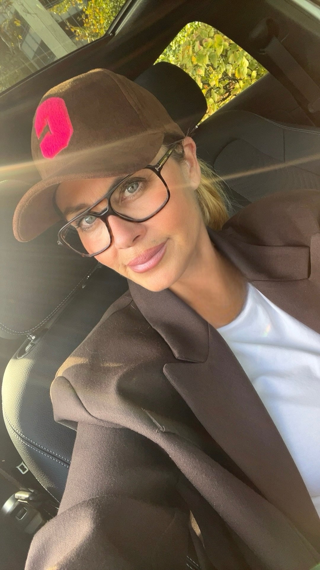 Frau mit brauner Velour Cap mit pinkem C Logo, schwarzer Brille und brauner Blazer sitzend im Auto.