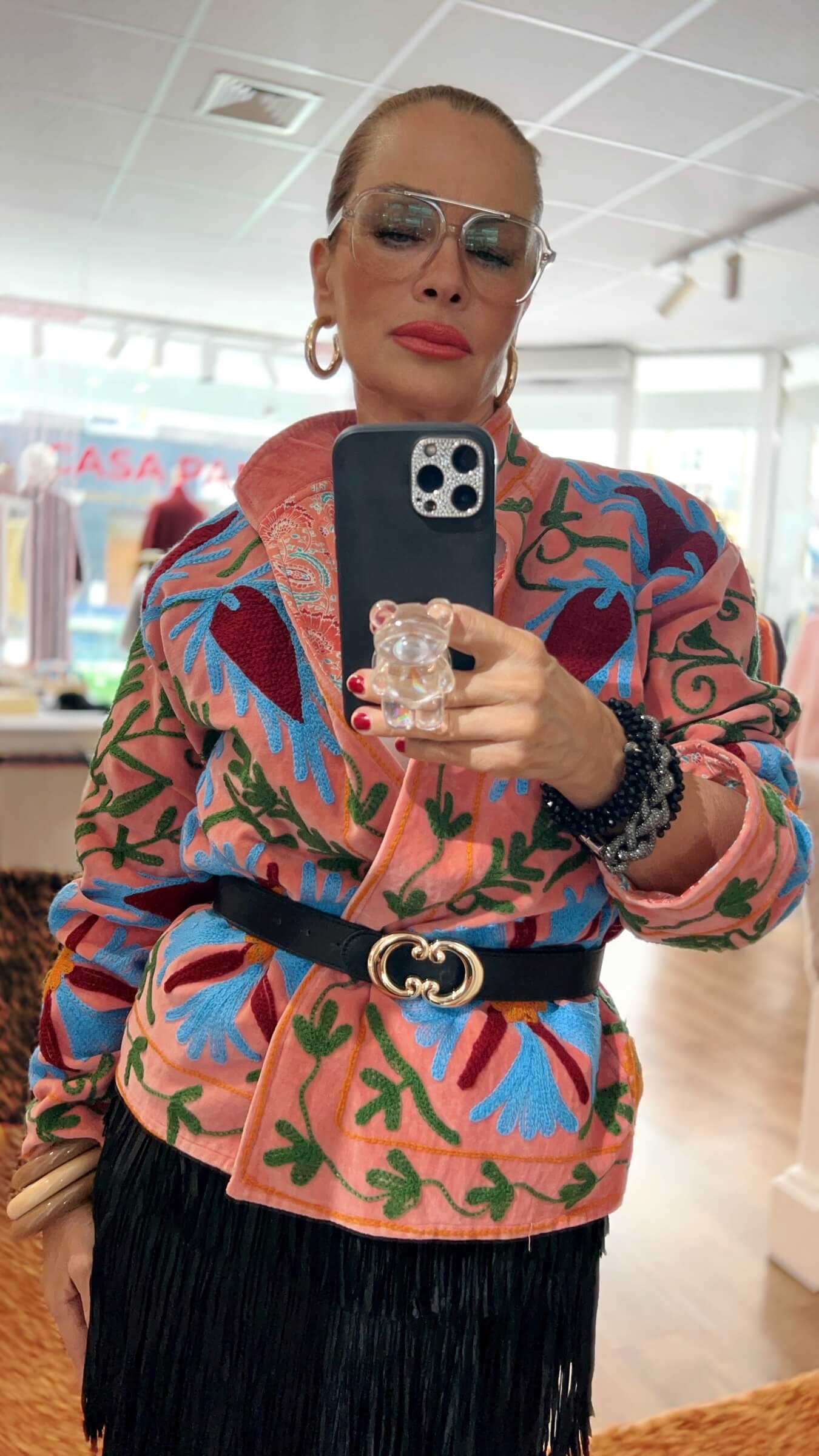 Nahaufnahme einer Lachs rosafarbenen, floral bestickten Jacke mit blau-grünen Stickereien, kombiniert mit schwarzem Gürtel und schwarzem Fransenrock in einer hellen Boutique.