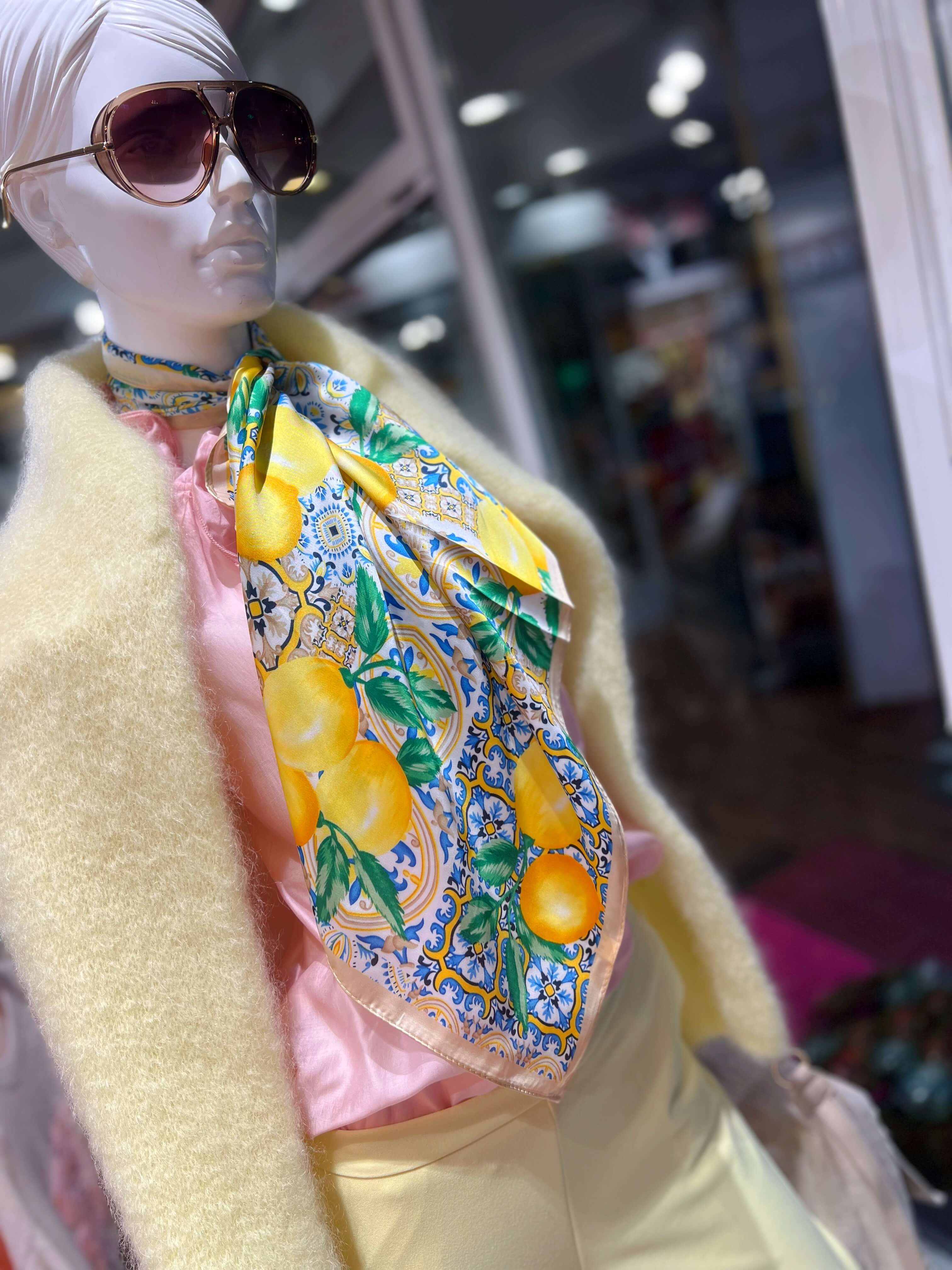 Schaufensterpuppe mit Sonnenbrille trägt ein quadratisches Tuch mit gelbem Zitronenmuster und blauem Kachelmotiv, kombiniert mit hellem Pullover und heller Hose vor Schaufenster.