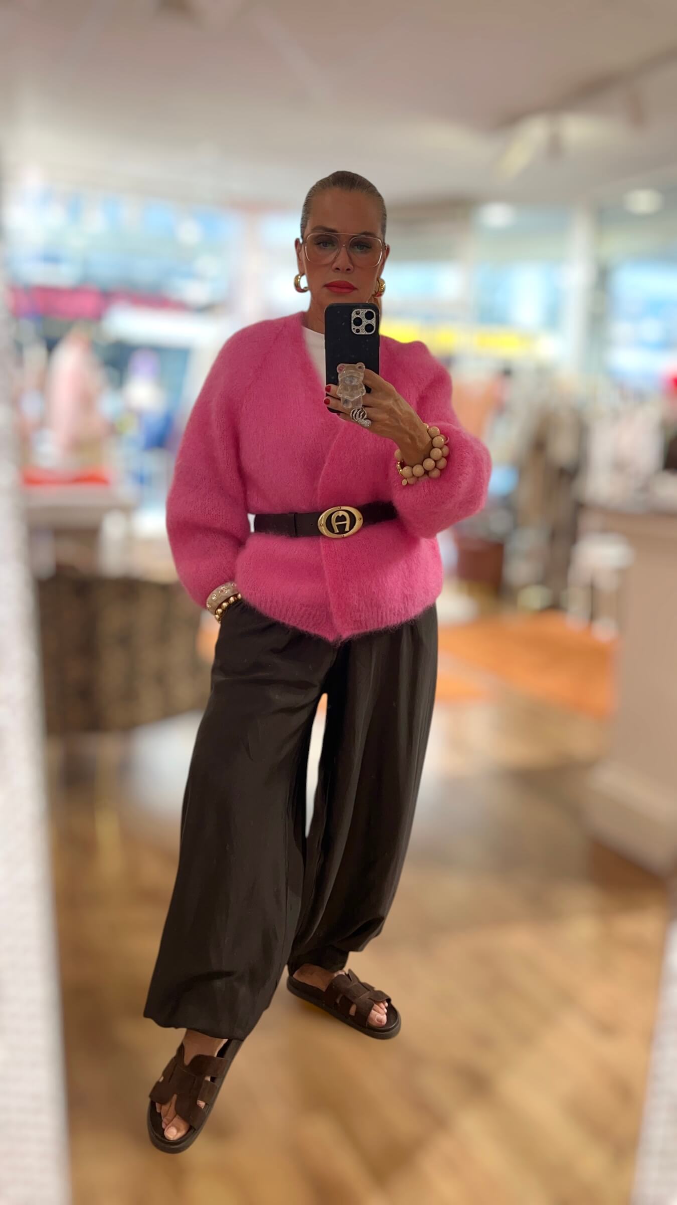 Frau trägt eine flauschige pinke Kid-Mohair-Strickjacke mit Gürtel, weite schwarze Hose und Sandalen und steht vor einem hellen Boutique-Hintergrund.