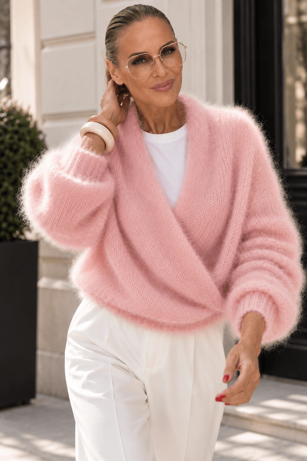 Frau trägt eine weiße Hose, weißes T-Shirt und eine rosa farbenfroh a Strickjacke und steht auf einer Straße