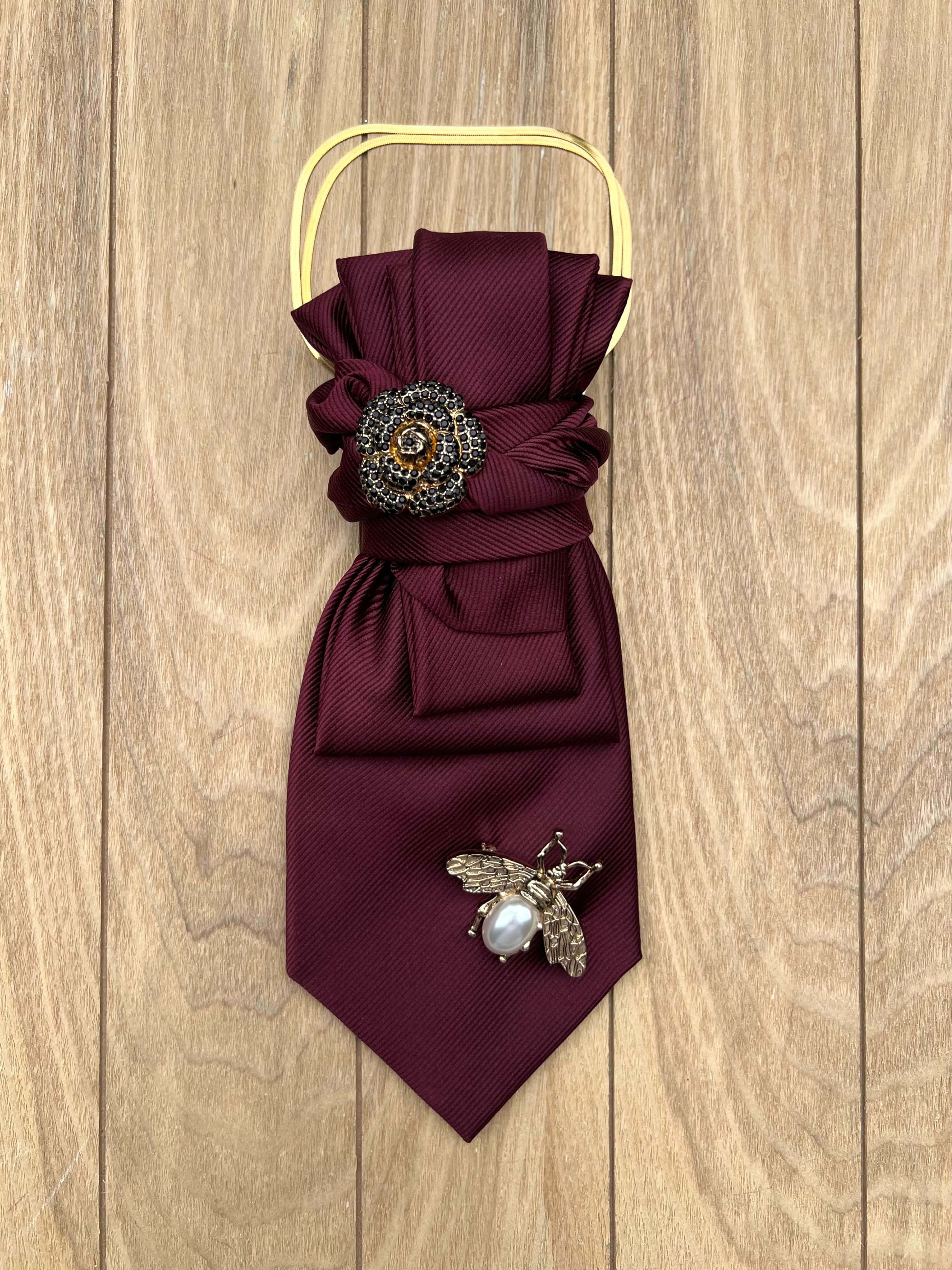 Bordeauxfarbene Statement-Krawatte von Casapari mit goldfarbener Kette, floraler Brosche und Bienenbrosche auf Holzboden.