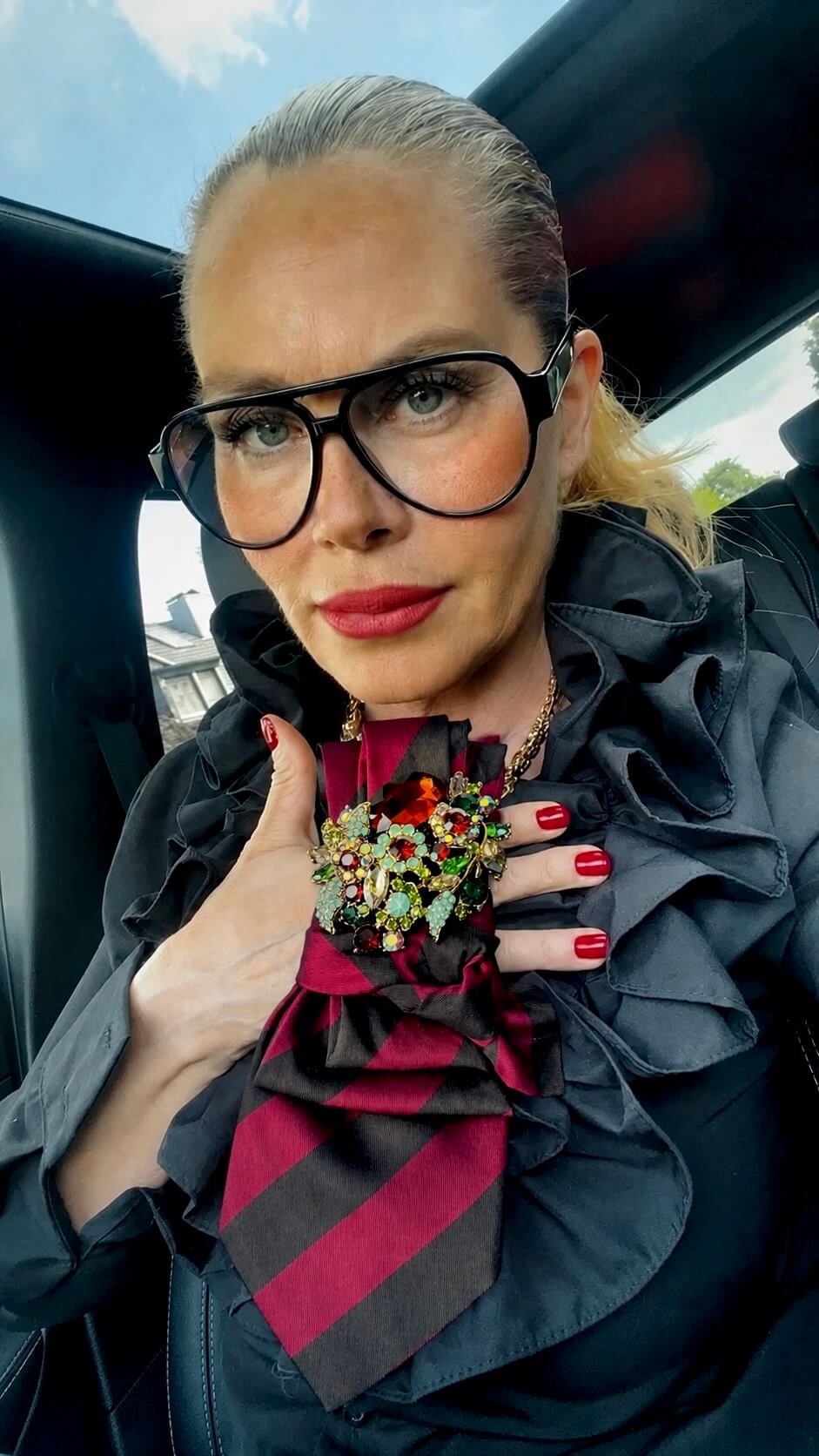 Frau trägt schwarze Bluse mit gestreift Krawatte und Brosche und großer schwarze Brille im Auto fotografiert