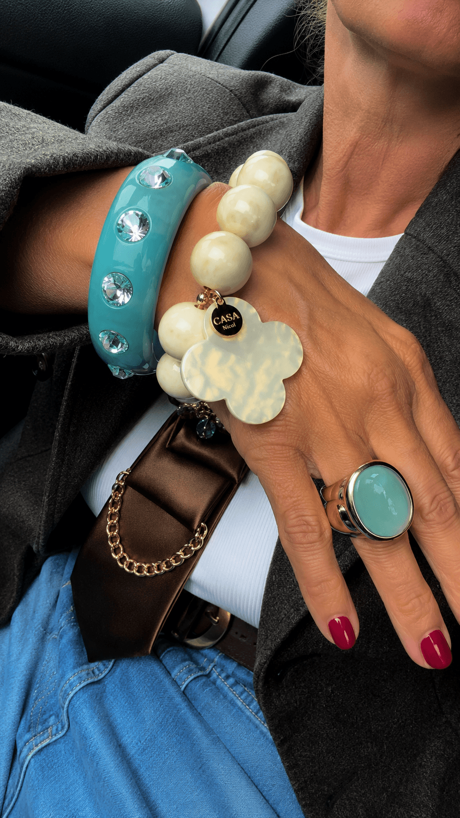 Handgelenk mit blauen Resina-Armreif mit Strass-Steinen, kombiniert mit einem cremefarbenen Perlenarmband und einem blauen  Ring. Die Person trägt einen grauen Blazer, Jeans und eine braune Krawatte mit goldfarbener Kette.