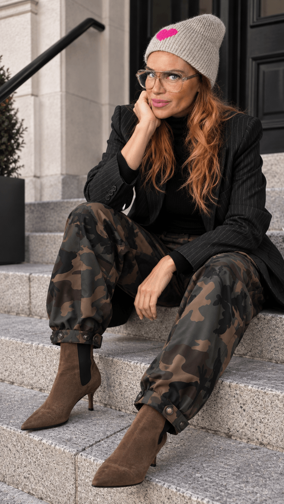 Frau sitzt auf einer Steintreppe und trägt die Barrel Pants Urban Camo mit schwarzem Blazer, braunen Stiefeletten und beiger Mütze.