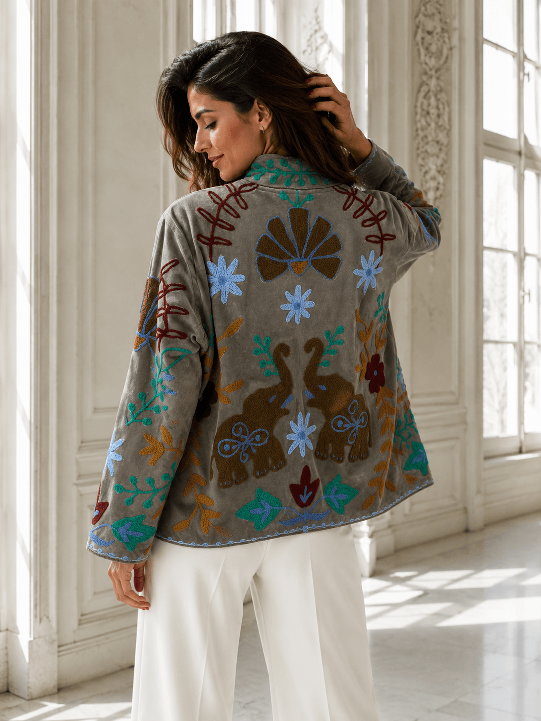 Frau in heller Hose trägt eine bestickte Jacke mit floralen Details und Elefantenmotiven in einer hellen Innenraumkulisse, Rückenansicht