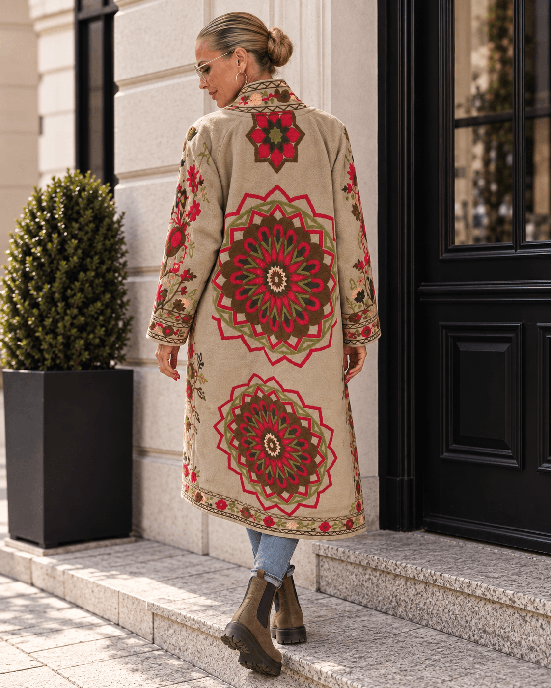 Frau in Skinny Jeans und braunen Chelsea Boots trägt einen langen beigefarbenen Mantel mit großen floralen Stickereien auf dem Rücken.