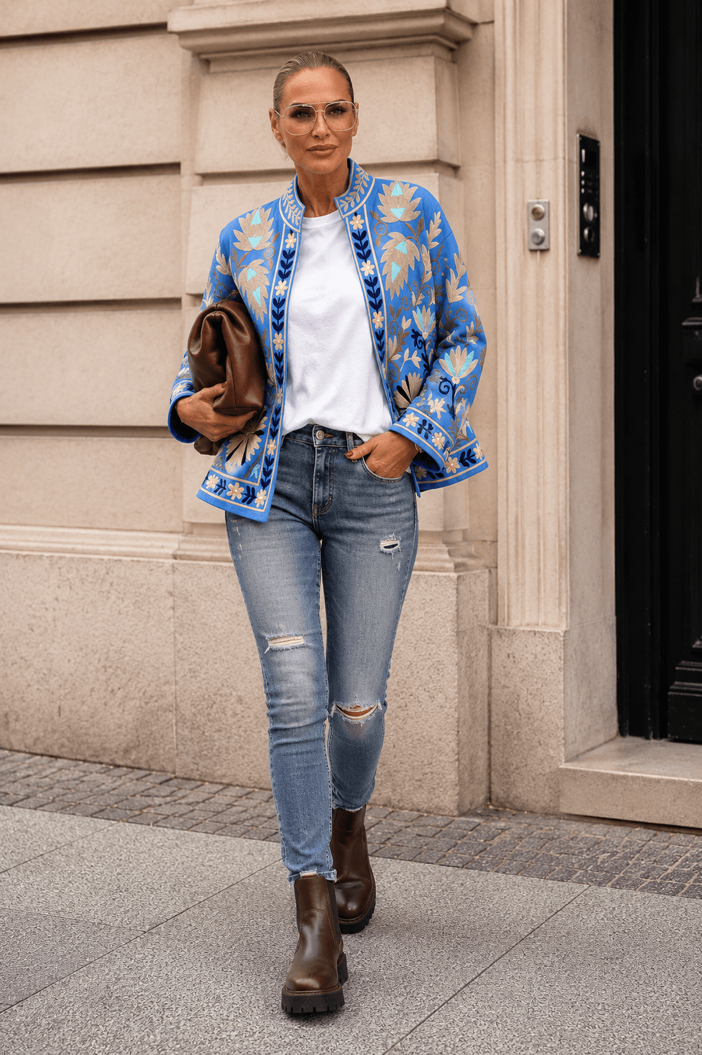 Frau in blauer bestickter Jacke über weißem T-Shirt, Skinny Jeans und braunen Chelsea Boots vor heller Häuserfassade.