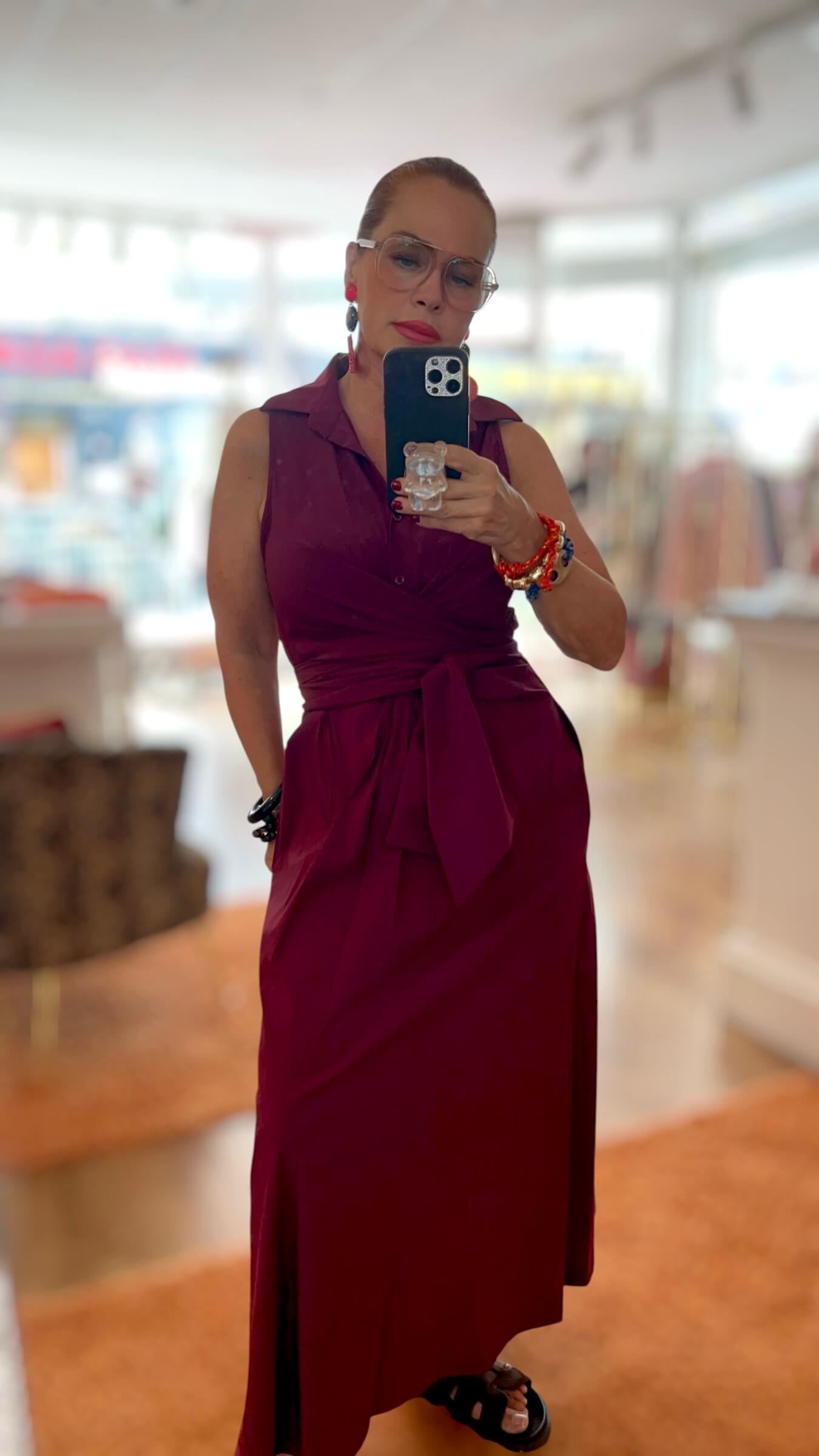 Frau trägt ein bordeauxrotes Wickelkleid mit Bindegürtel und Knopfleiste. Das ärmellose Kleid wird in der Taille gebunden und fällt fließend bis zum Knöchel. Kombiniert mit einer dekorativen Brosche am Ausschnitt und farbigen Armbändern.