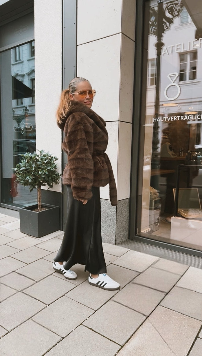 Frau mit Brille trägt ein braunes Plüsch Jäckchen mit Bindegürtel, weiten Rock und weiße Sneaker. Sie steht vor einem Schaufenster in der Stadt.