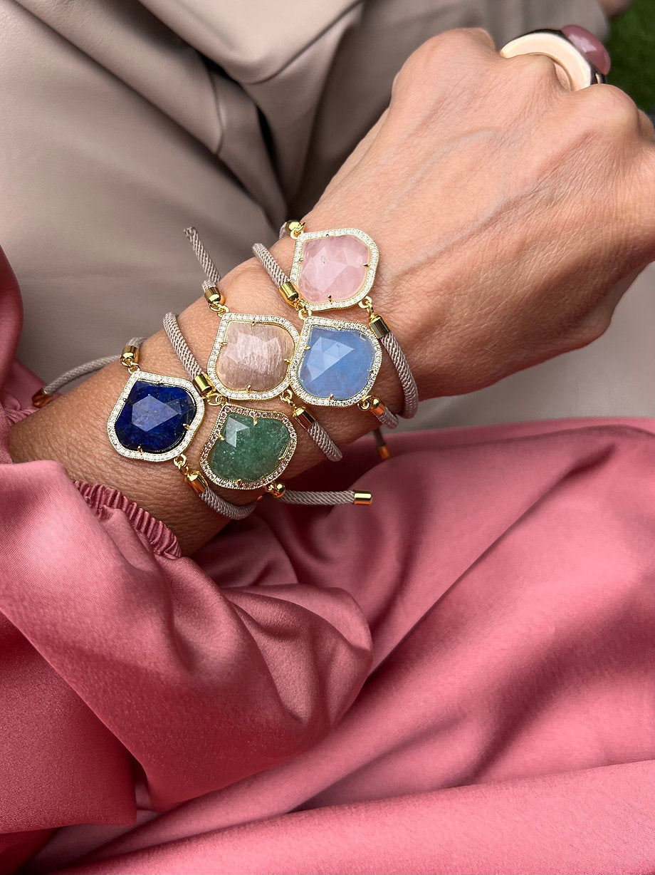 Fünf handgefertigte Armbänder aus der Glow & Grace Kollektion mit funkelnden Steinelementen in sanften Rosé, Grau und Goldtönen, locker arrangiert auf roséfarbenen Stoff.