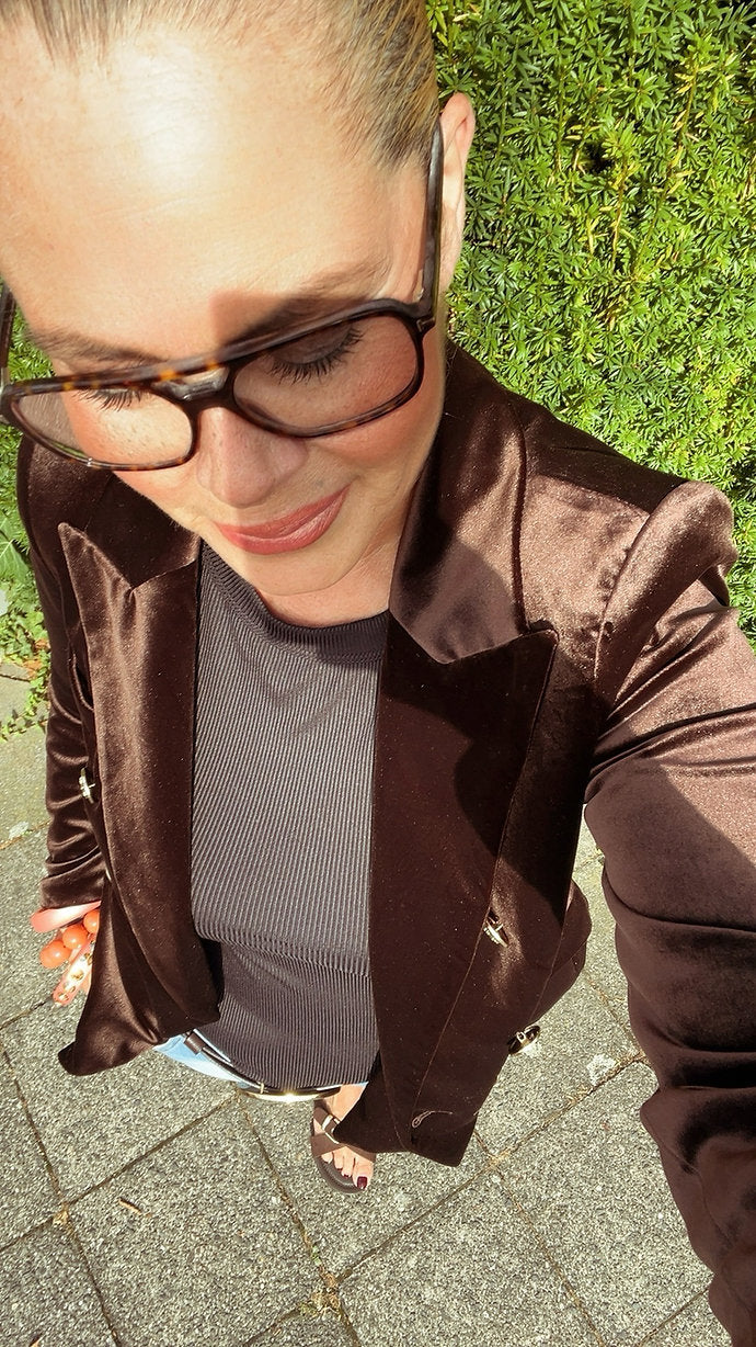 Frau mit hochgesteckten Haaren trägt einen schokobraunen Samtblazer, Brille und ein graues Oberteil. Selfie von oben, draußen bei Tageslicht auf Steinboden.