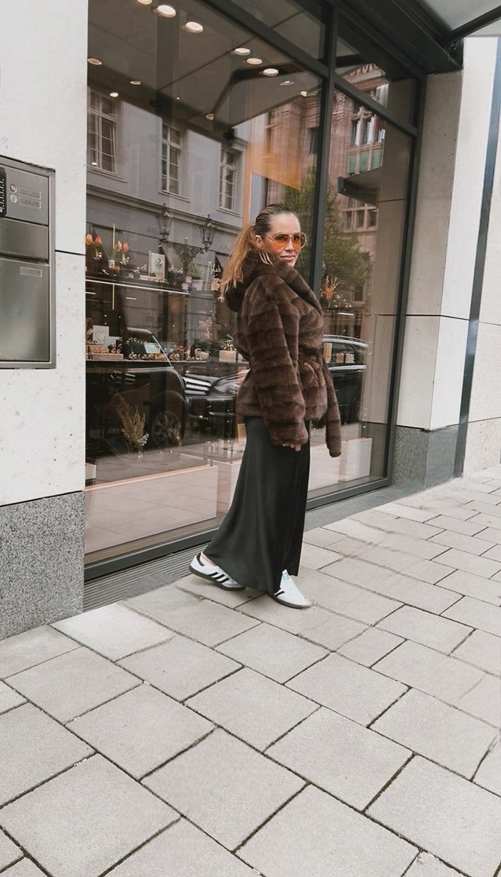 Frau mit Sonnenbrille trägt ein braunes Plüsch Jäckchen mit Bindegürtel, weiten Rock und weiße Sneaker. Sie steht vor einem Schaufenster in der Stadt.