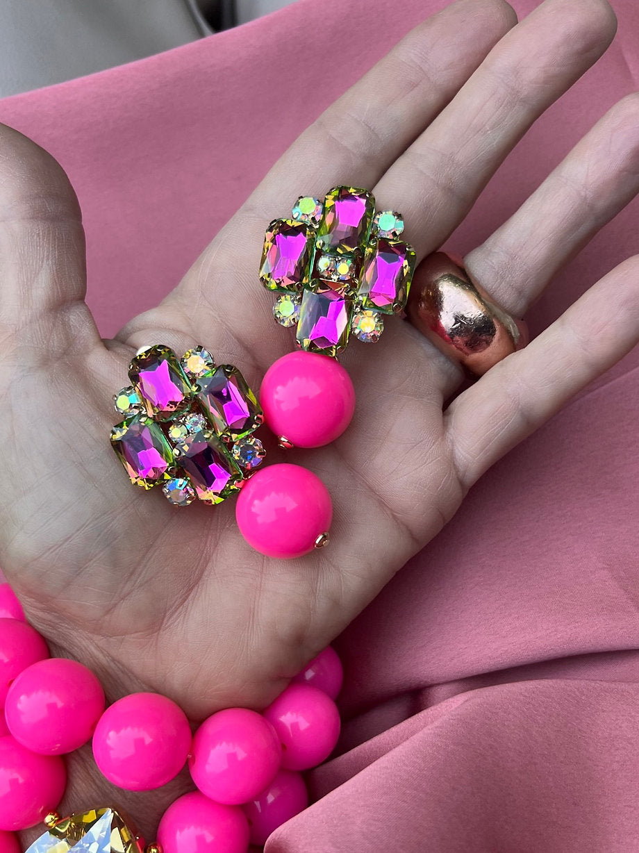 Hand mit goldfarbenem Ring hält ein Paar bunte Statement Clip Ohrringe mit pinken Kugeln und facettierten Kristallen in Violett und Grün. Im Hintergrund liegt ein Armband mit großen pinken Perlen auf roséfarbenen Stoff.