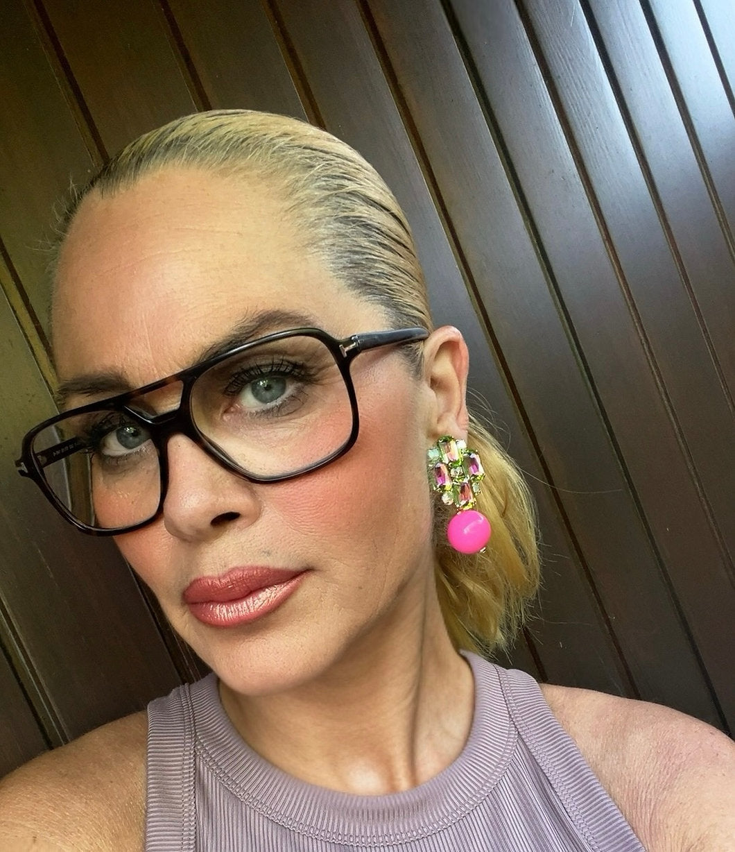 Frau mit blondem, streng zurückgebundenem Haar trägt große, bunte Statement Clip Ohrringe in Pink, Grün und Violett. Sie trägt eine Brille und ein ärmelloses Oberteil.