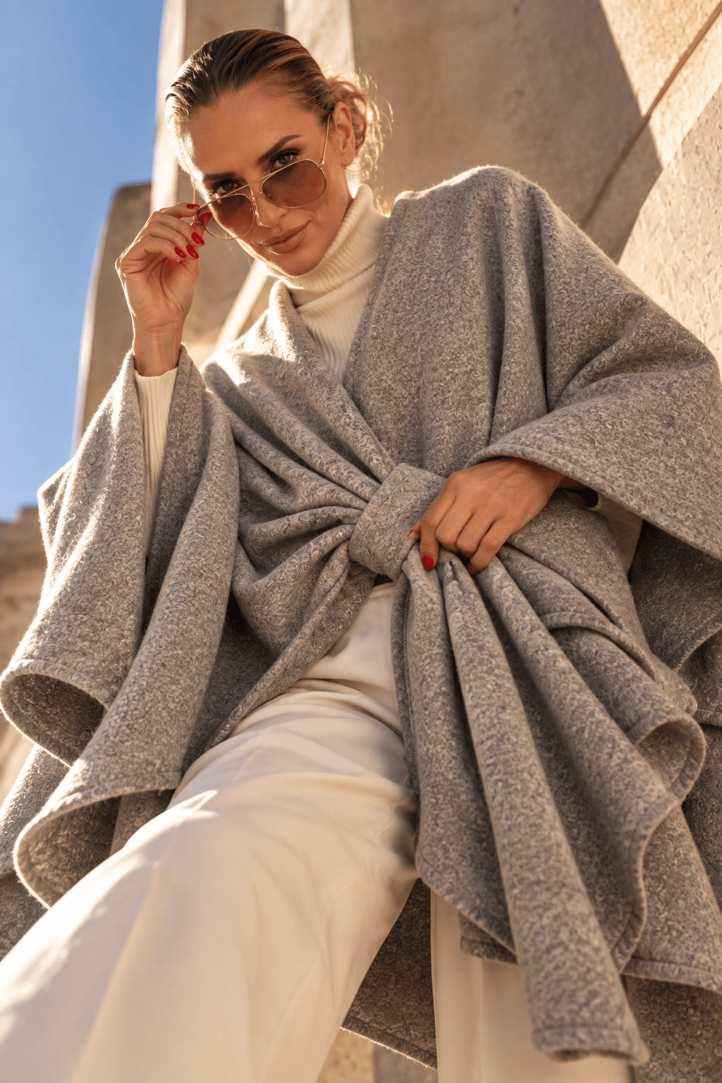 Frau trägt ein taupefarbenes Cape mit Drapierung vorne über einem cremefarbenen Rollkragenpullover und heller Hose