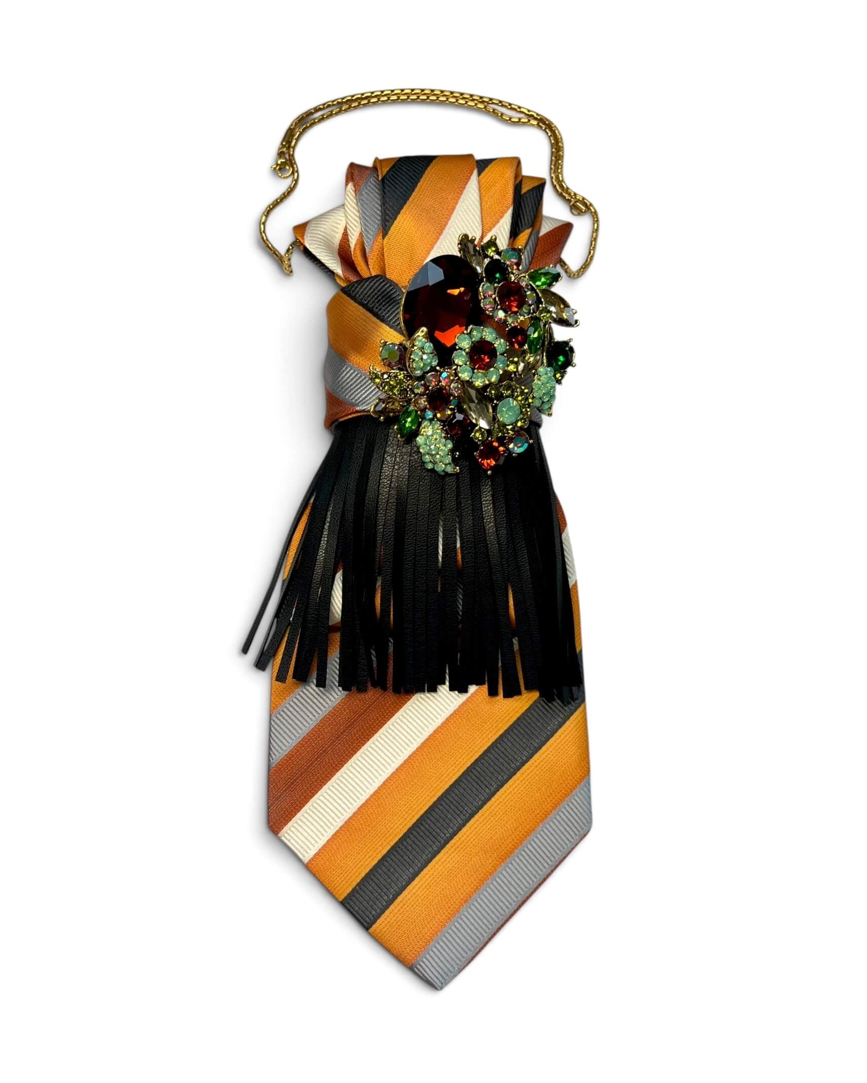 Gestreifte Krawatte in Orange, Creme und Grau mit schwarzen Lederfransen, mehrfarbiger Schmuckbrosche und goldfarbener Kette auf hellem Hintergrund.