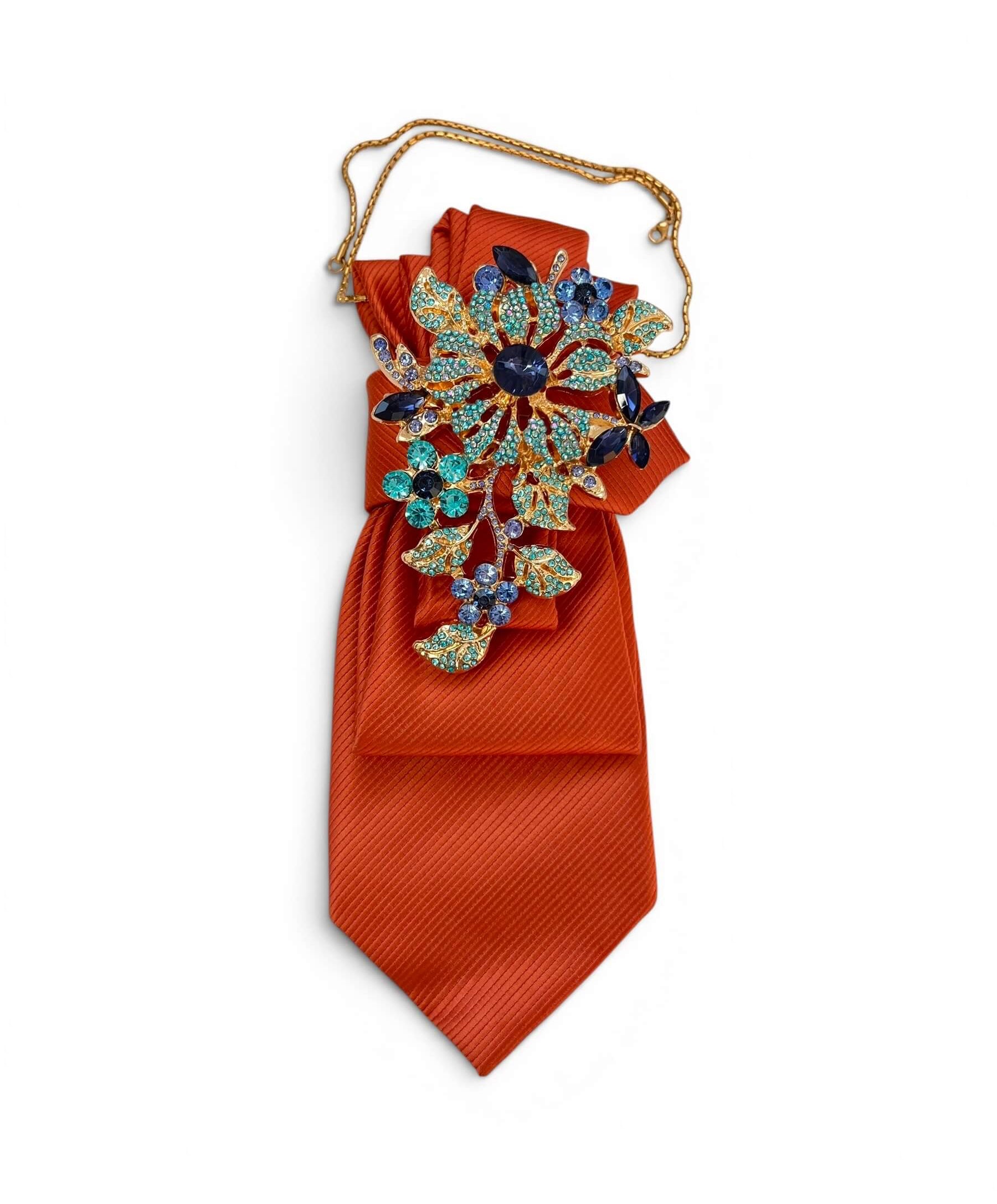 Orangefarbene Krawatte mit goldfarbener Kette und großer Brosche mit blauen und türkisfarbenen Schmucksteinen auf hellem Hintergrund.